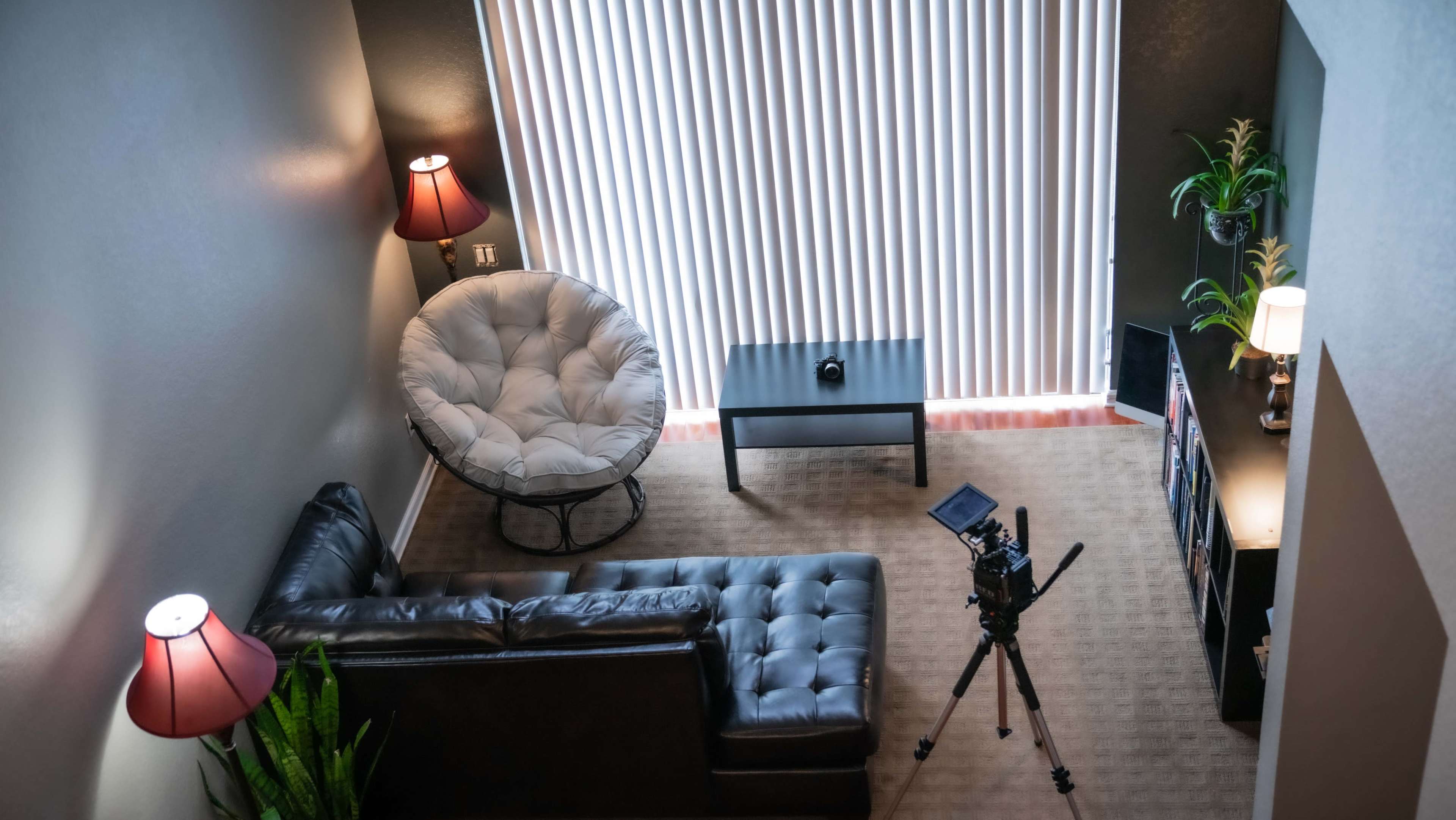A well-lit living room with a sectional sofa, a papasan chair, a coffee table, a camera set up on a tripod, and large vertical blinds allowing natural light to enter.