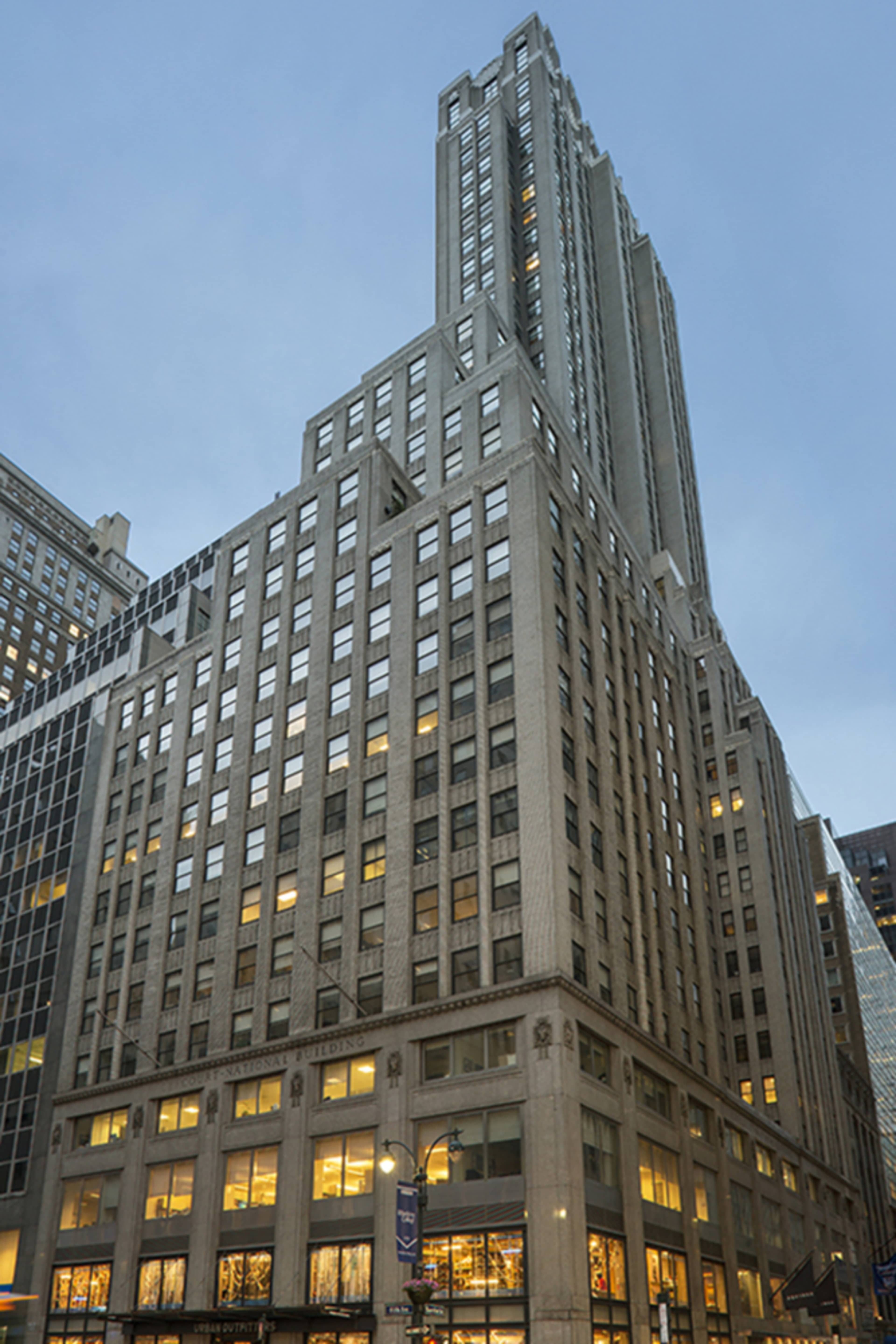 The image shows a tall skyscraper with a modern upper section and a decorated lower facade, illuminated at dusk.