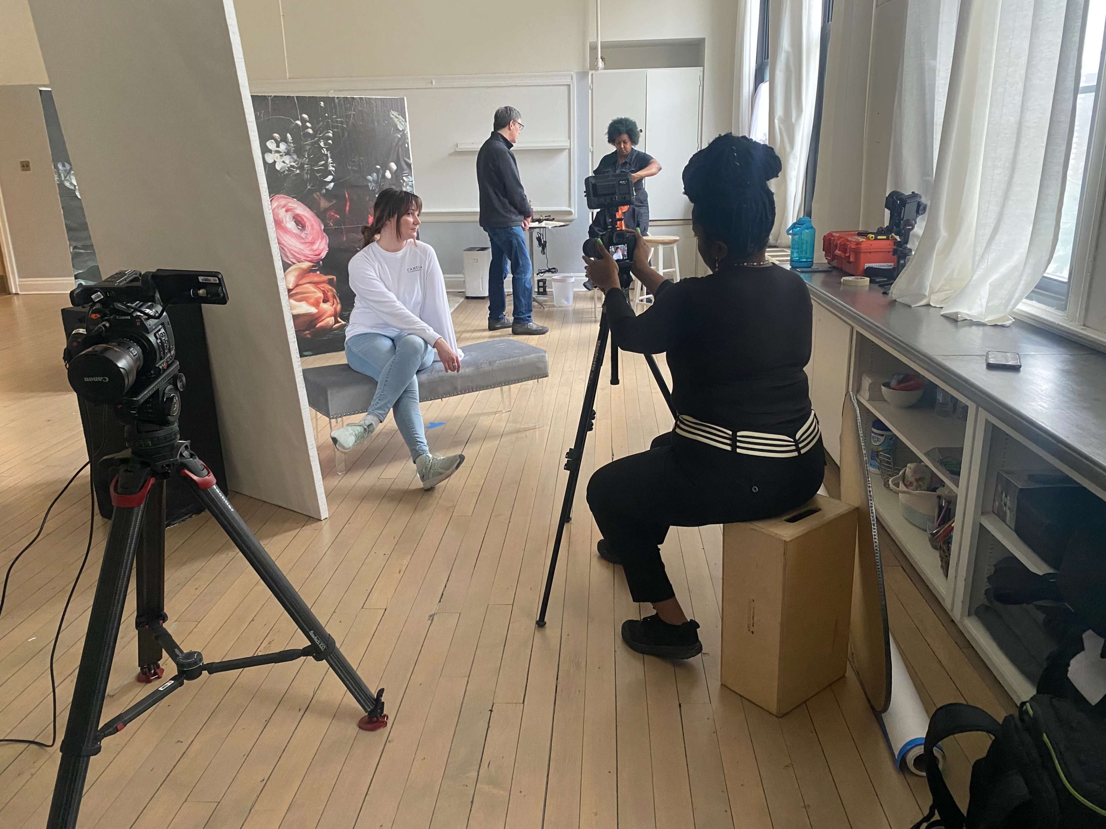 A woman sits in front of a camera while another person poses on a platform in a studio setting.