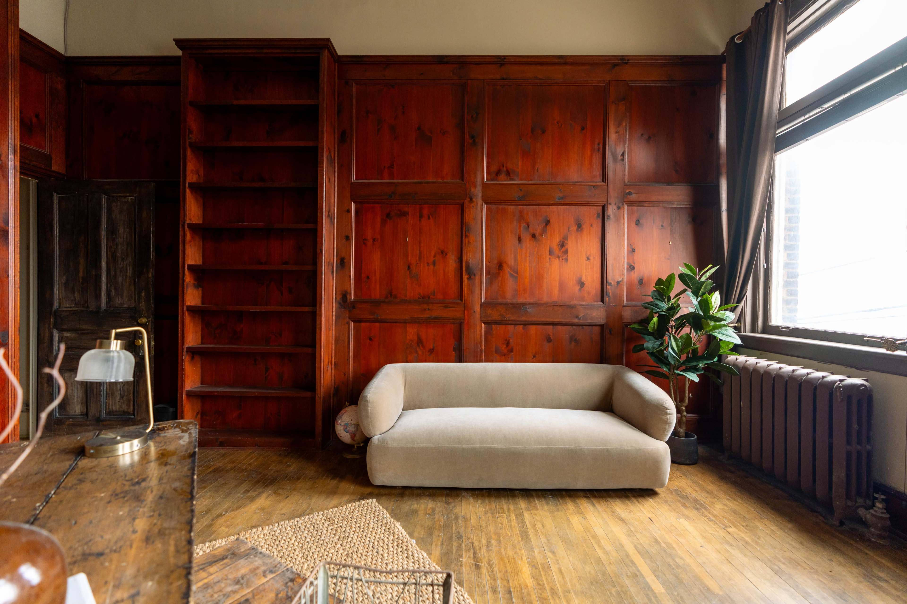 The image shows a room with wooden paneling on the walls, a beige couch, a bookshelf, a plant near a window, and hardwood flooring.
