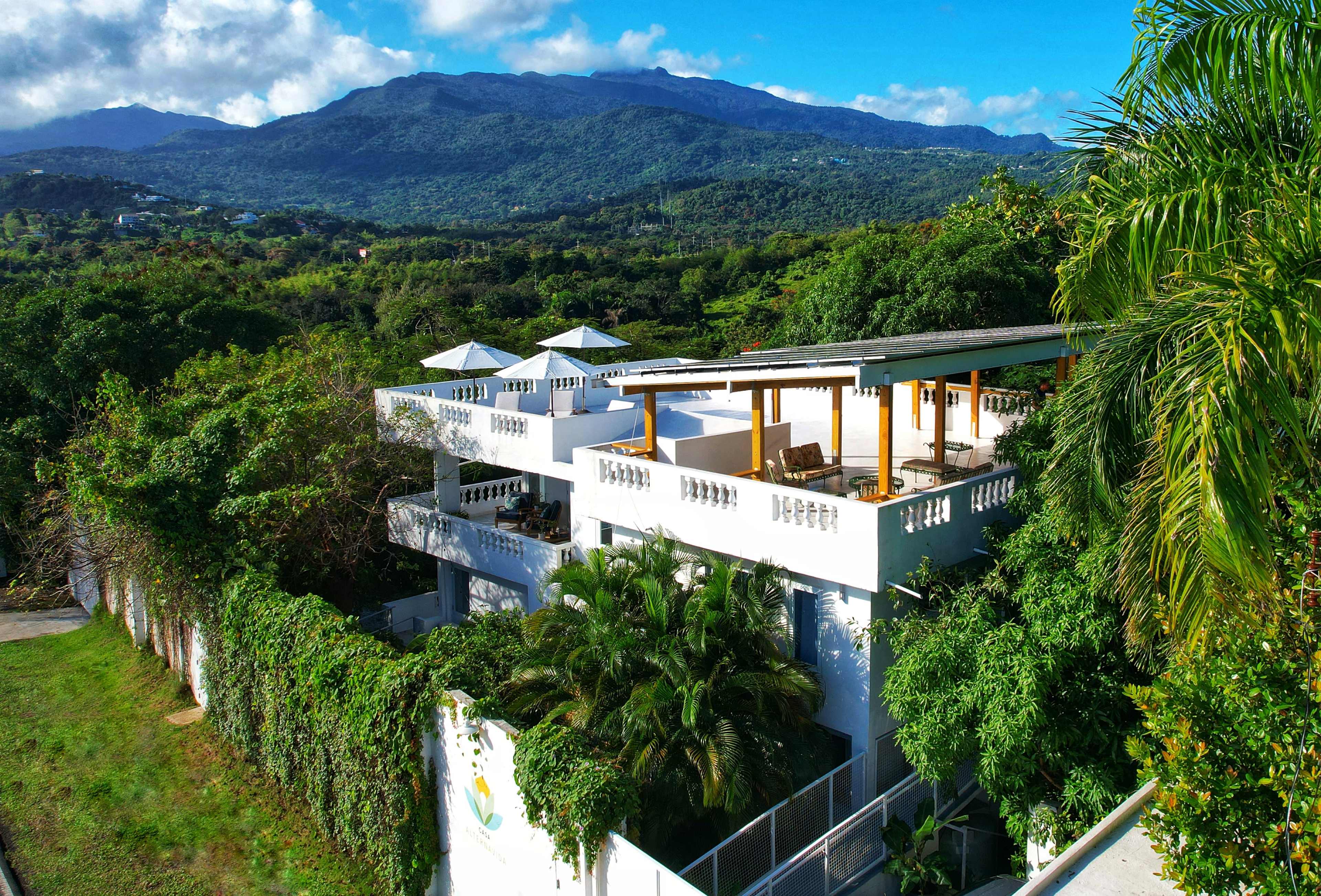 The image depicts a modern white house surrounded by lush greenery, with a mountainous backdrop under a partly cloudy sky.