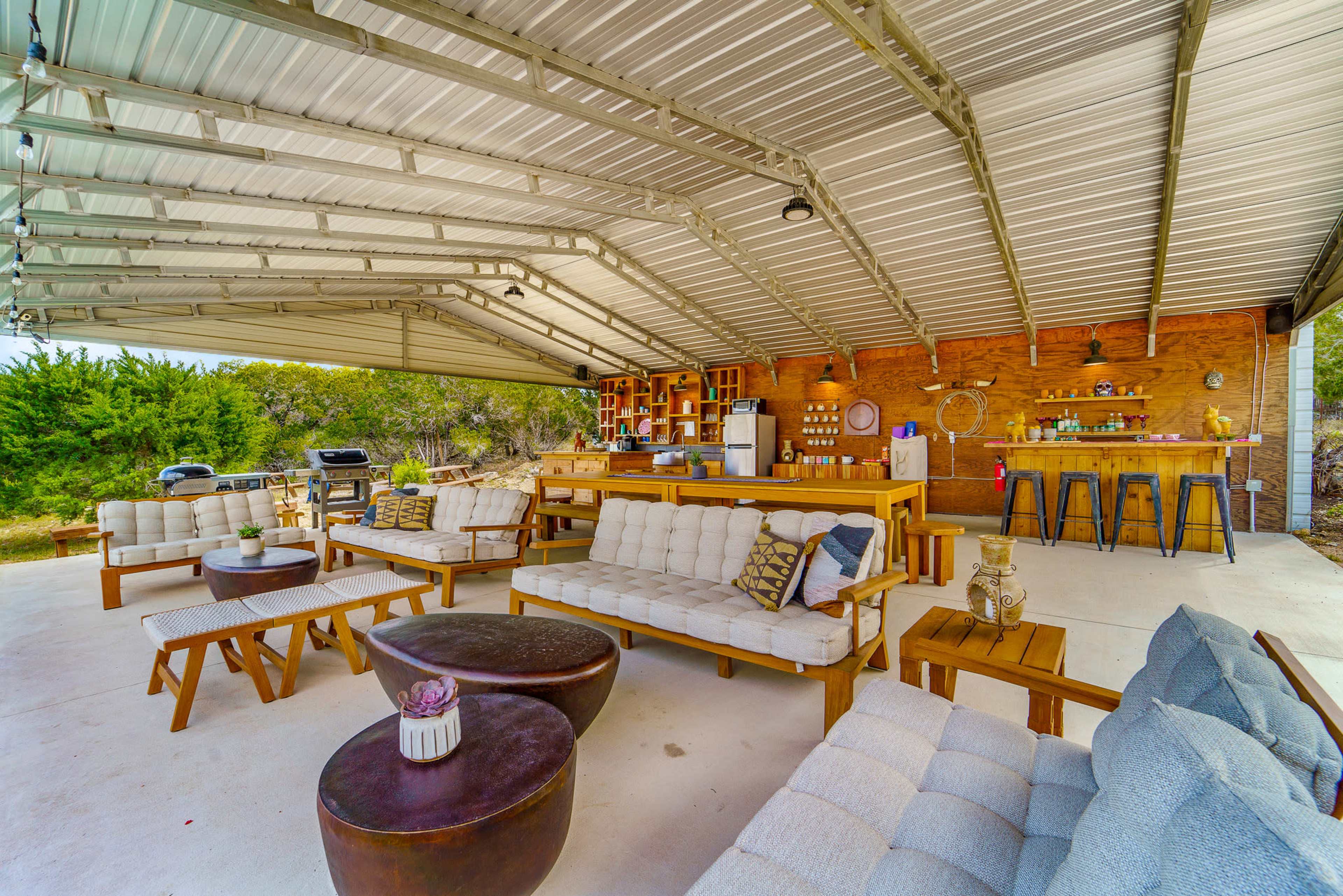 The image displays an outdoor lounge area with modern seating, wooden tables, and a kitchen bar under a metal roof structure surrounded by greenery.