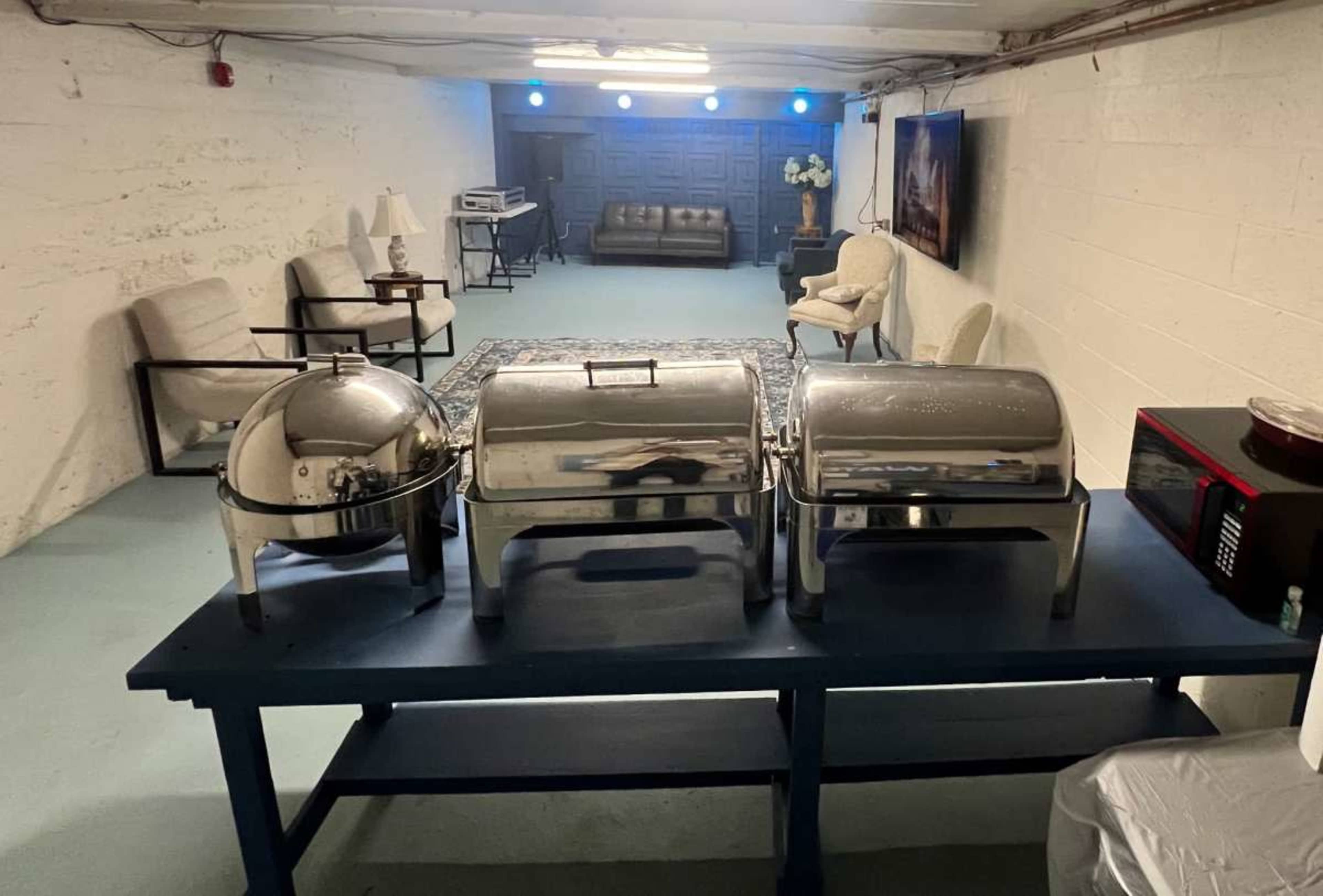 The image shows a basement space with a table displaying three silver food warming trays, and a cozy seating area visible in the background.