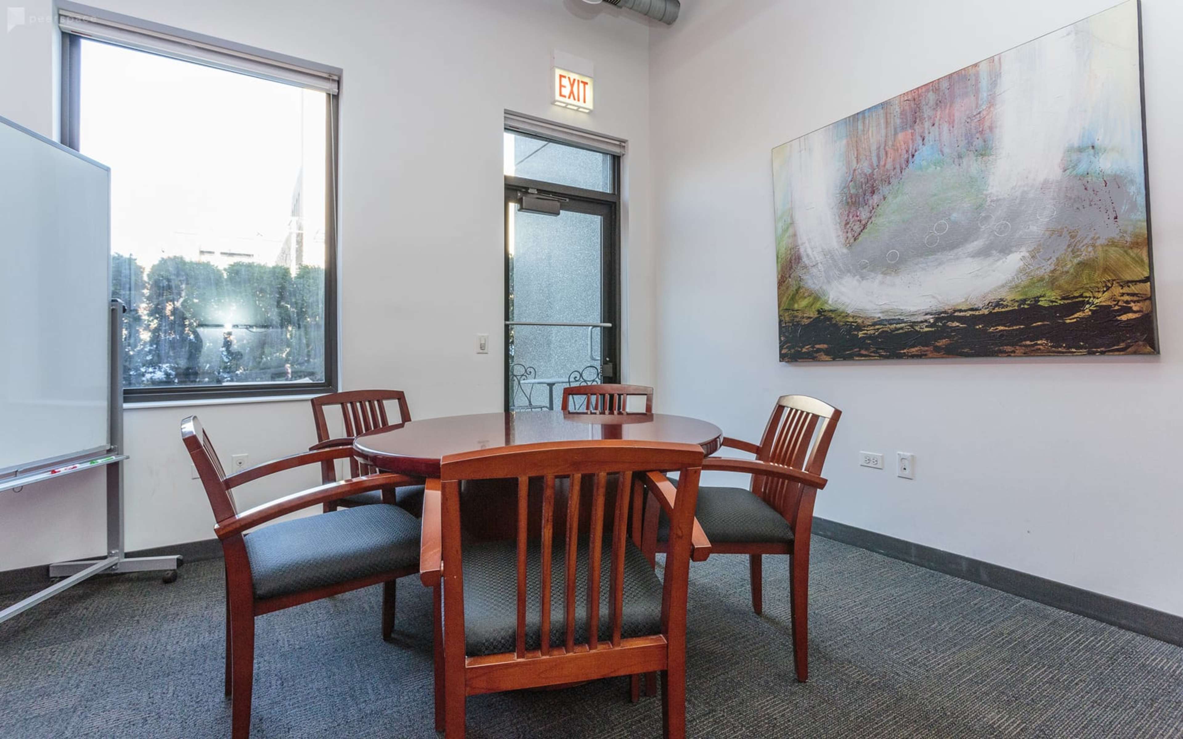 A meeting room features a round wooden table surrounded by four chairs, a whiteboard on wheels, and large windows letting in natural light.