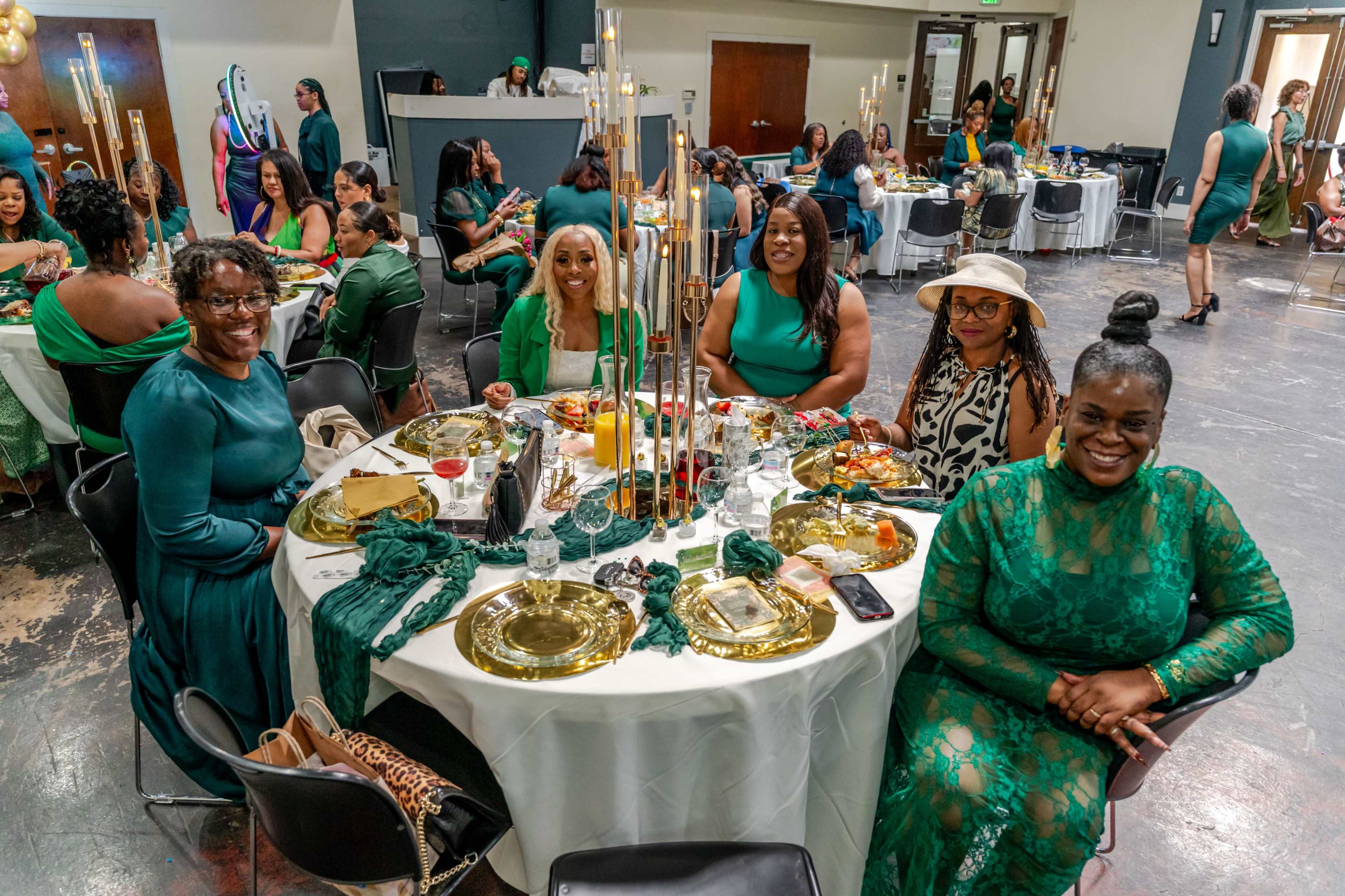 A group of women in green attire sits around a circular table set with plates and silverware, enjoying a meal at a social event.