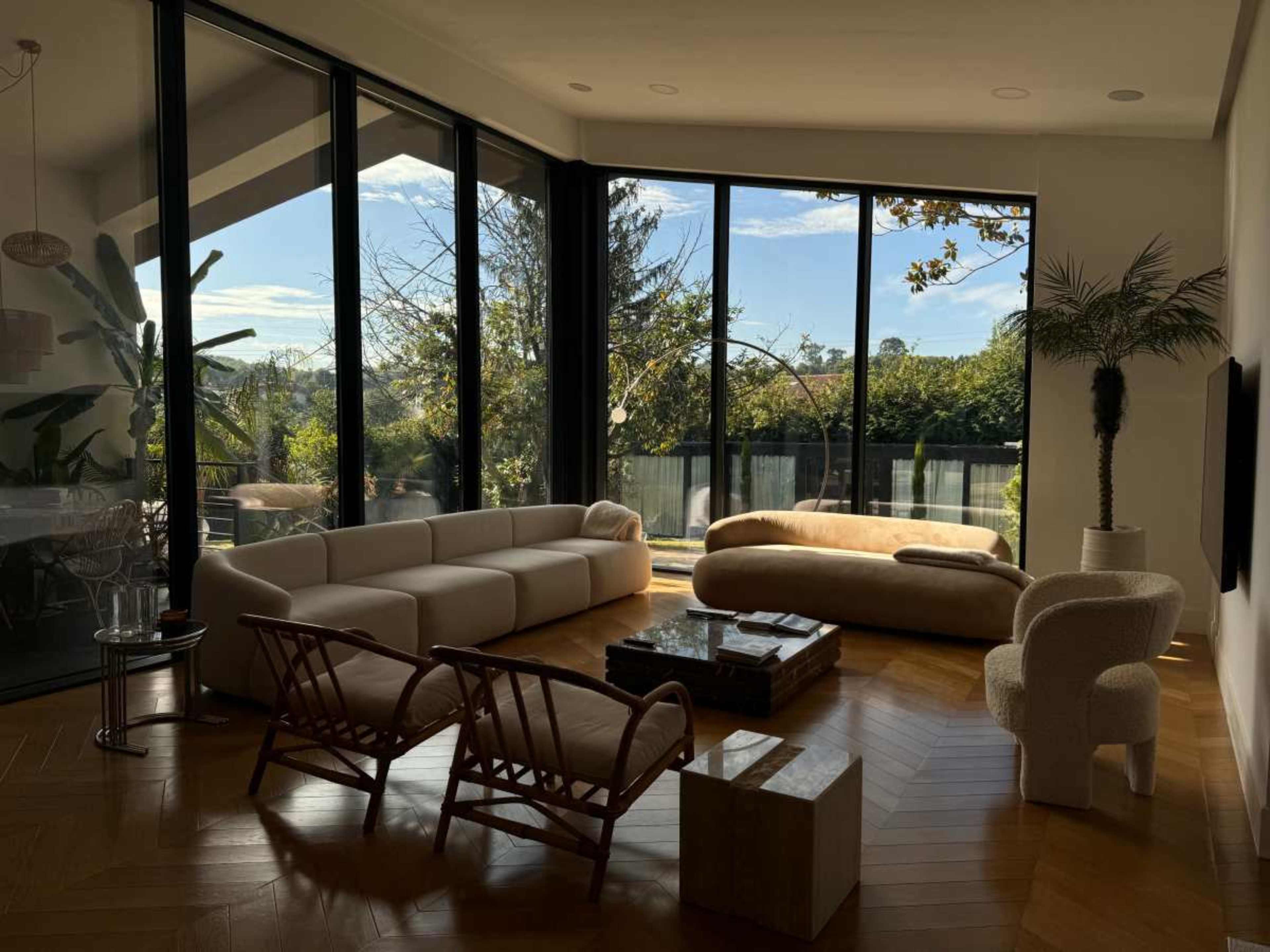 The image shows a modern living room with large windows, a curved sofa, a coffee table, and a view of greenery outside.