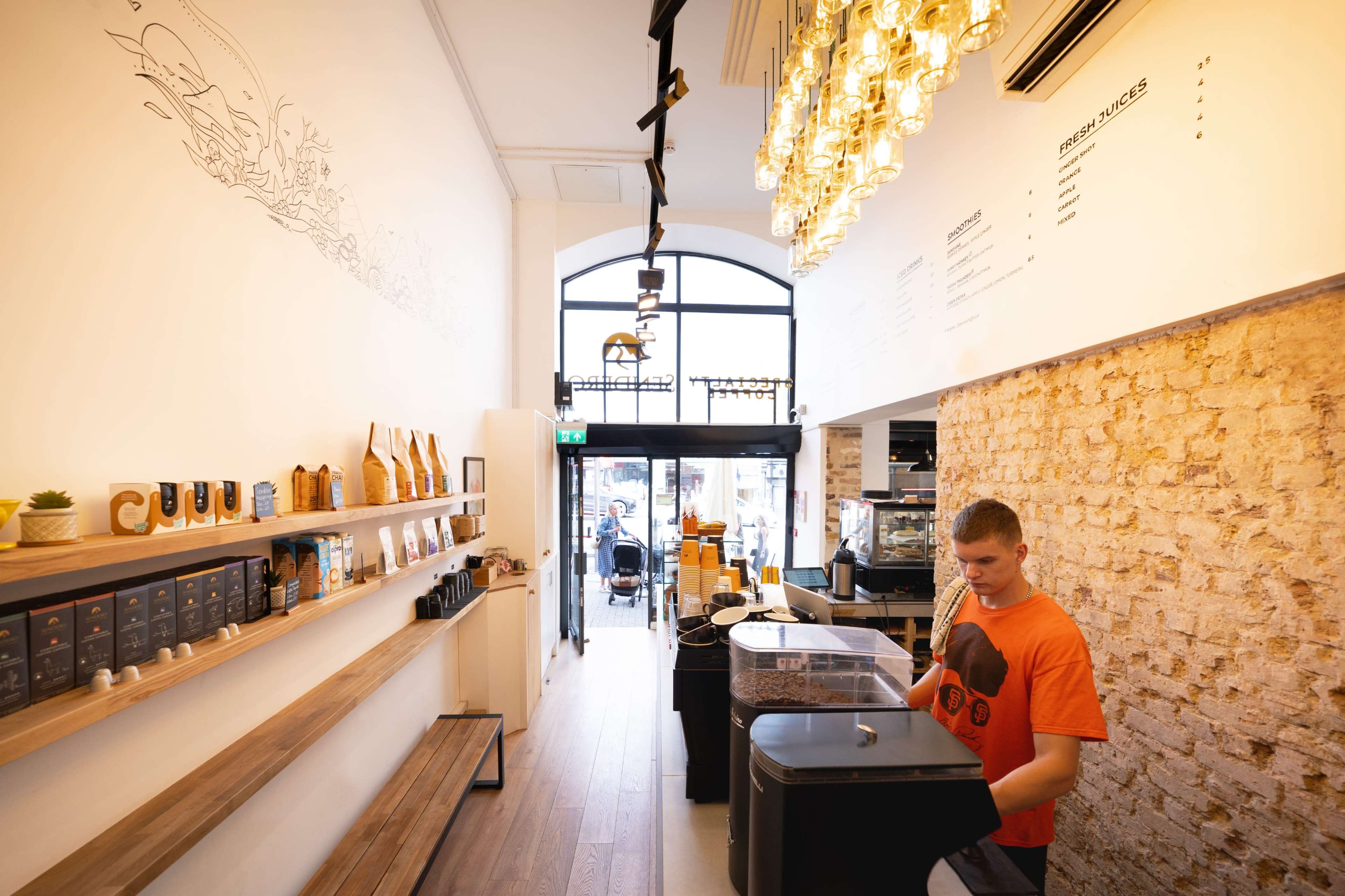 A barista prepares coffee inside a modern cafe with wooden shelves and a large window.