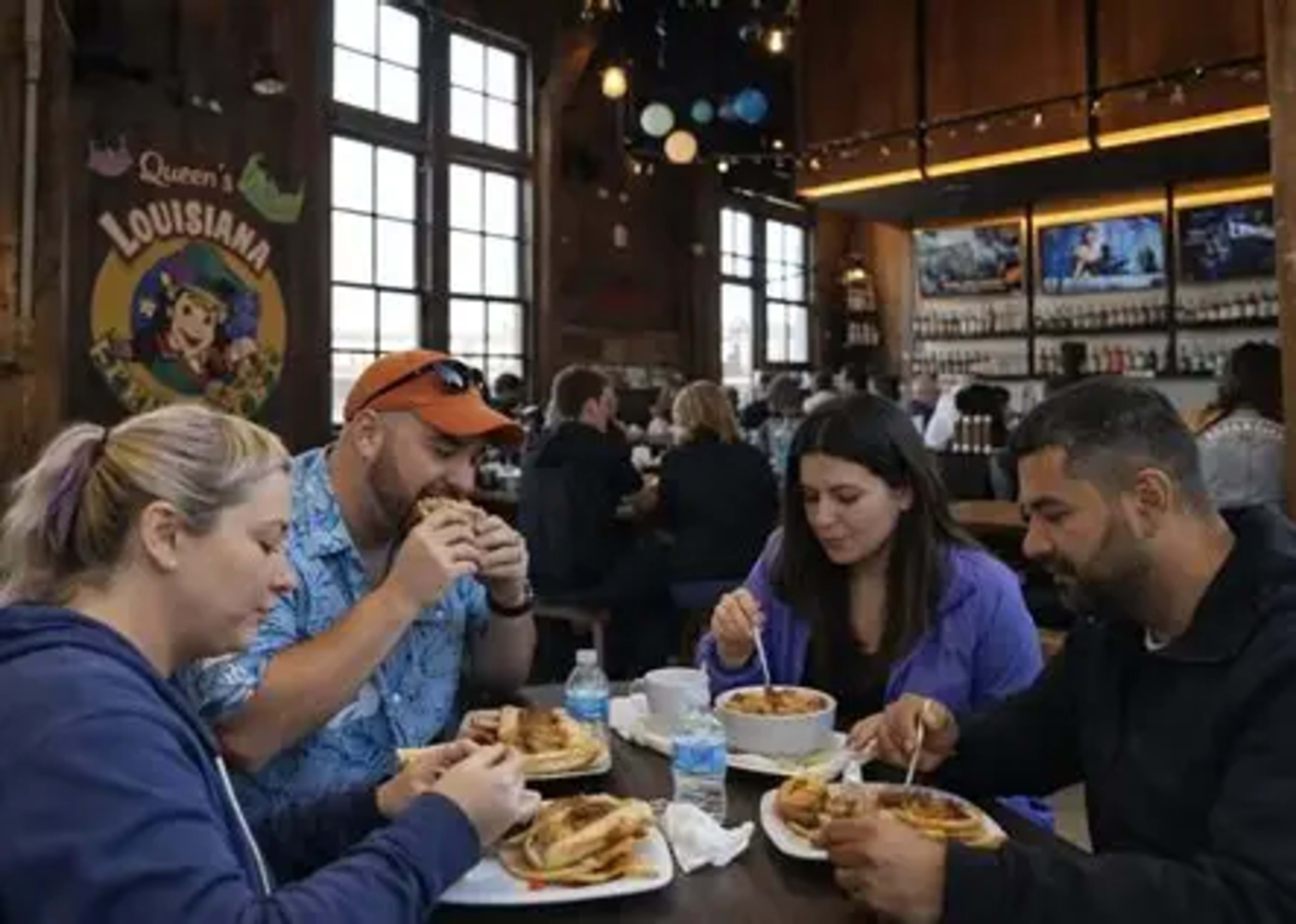Four people eat at a table in a lively restaurant with wooden decor and large windows.