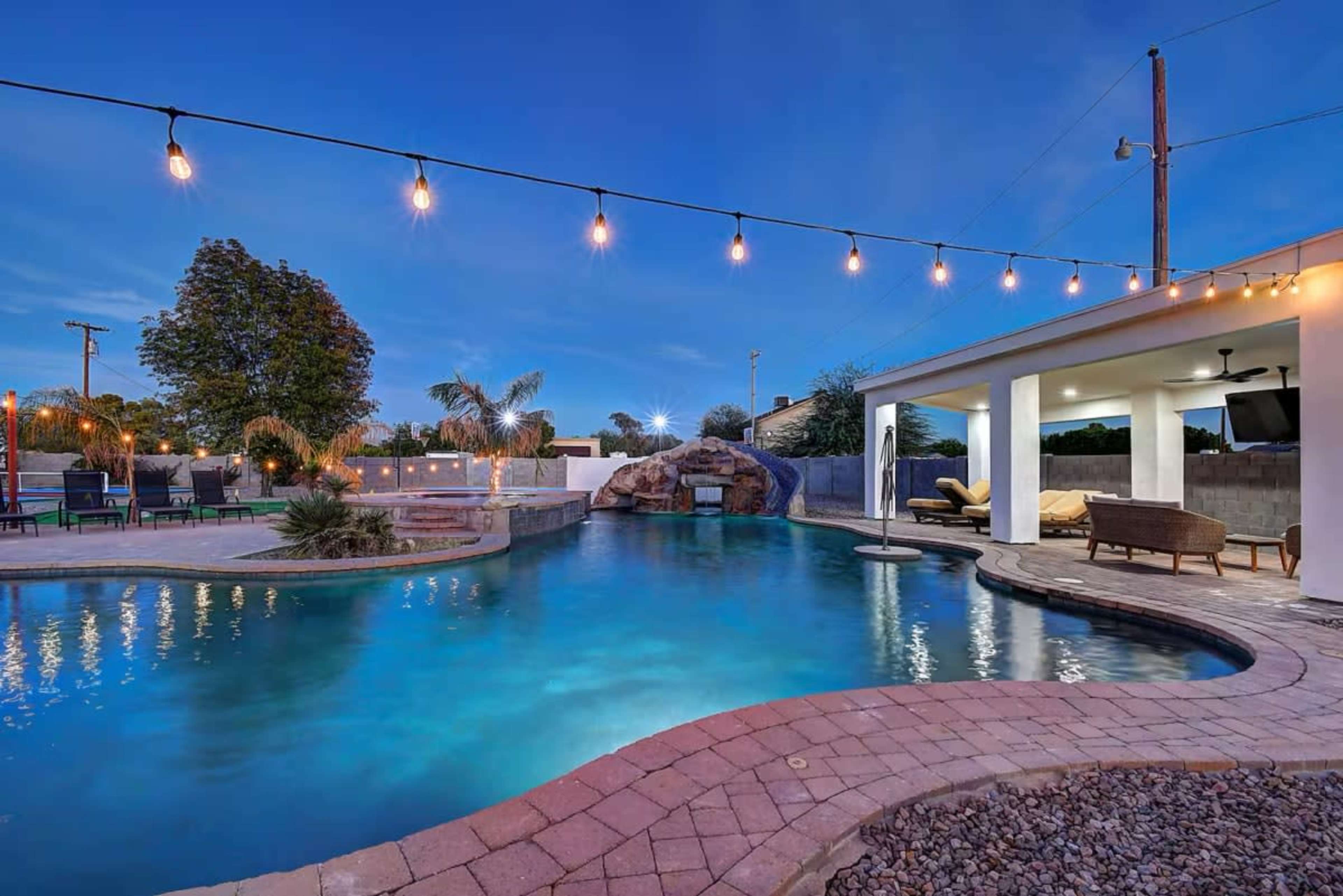 The image shows a swimming pool area with a rock waterfall feature, surrounded by landscaped plants, outdoor seating, and illuminated by string lights under a twilight sky.