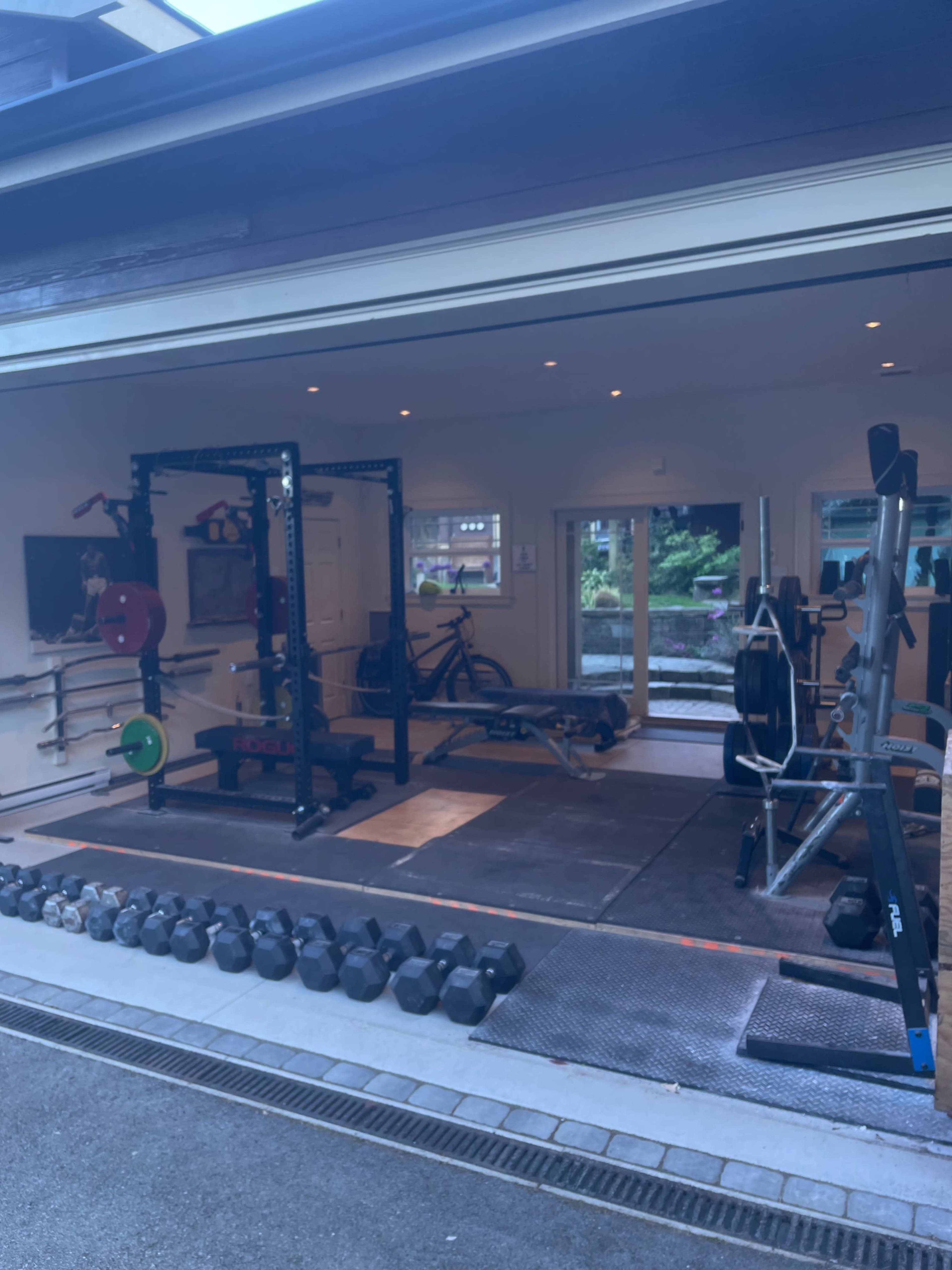 A home gym with various weightlifting equipment, including a squat rack, dumbbells, a bench, and exercise bikes, all arranged in a spacious garage setting.