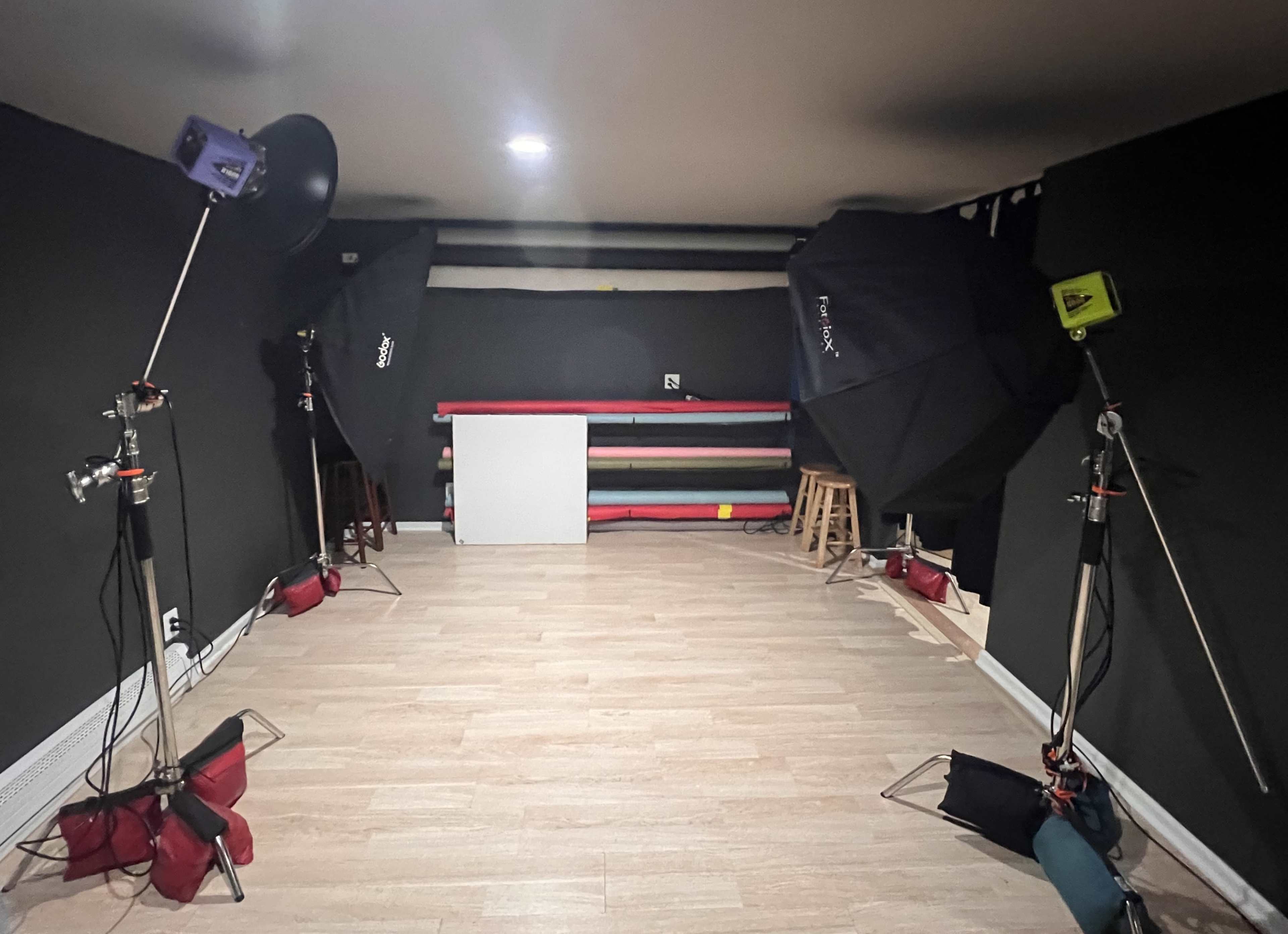 A photography studio with two softbox lights, a white backdrop, and a rack of colored paper rolls against a dark wall.