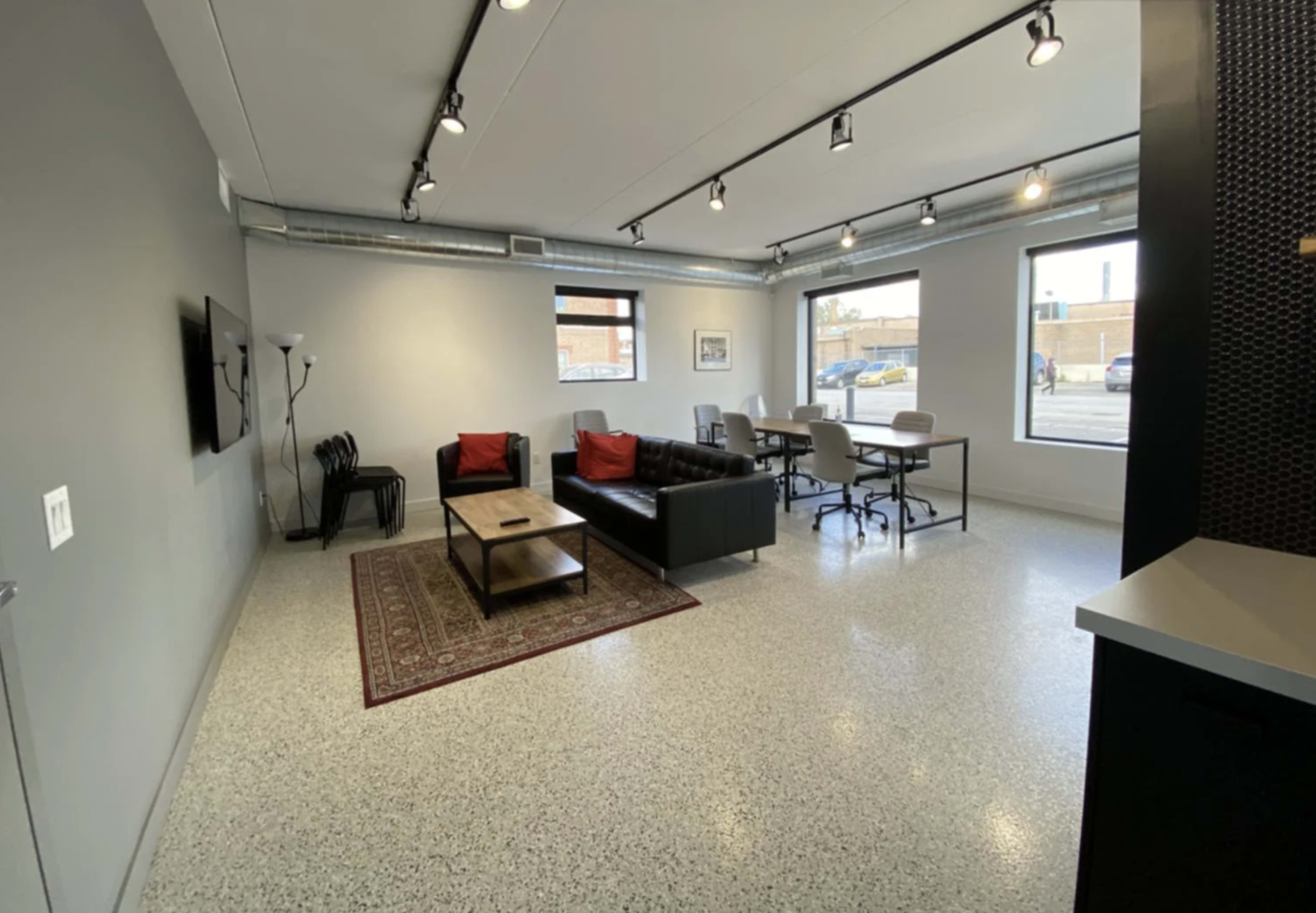 A modern office space with a seating area featuring a black leather sofa, a coffee table, and a dining area with white chairs and a large table, illuminated by overhead lighting.
