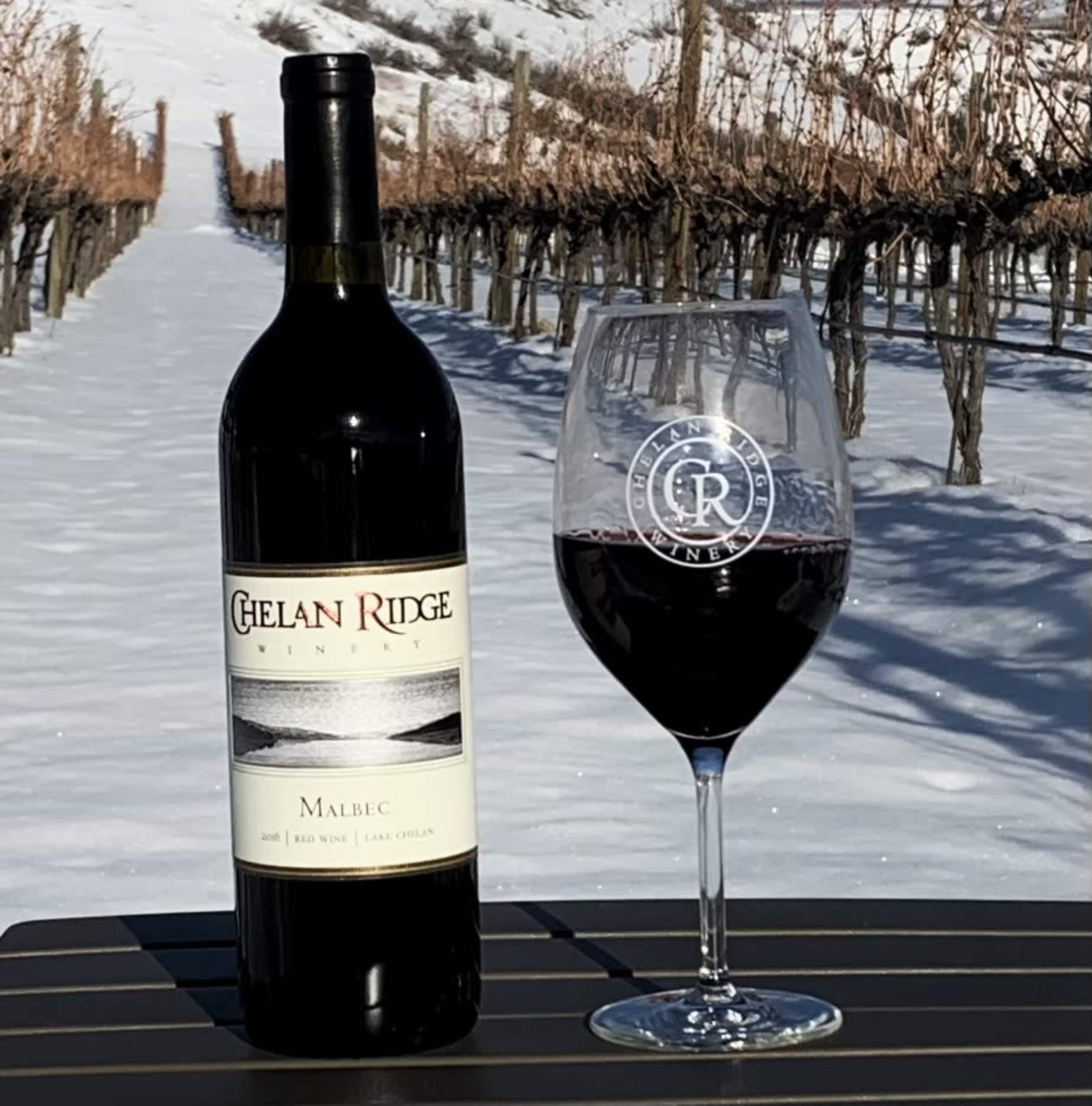 A bottle of Chelan Ridge Malbec wine stands next to a filled wine glass on a table, with vineyard rows and snow-covered ground in the background.