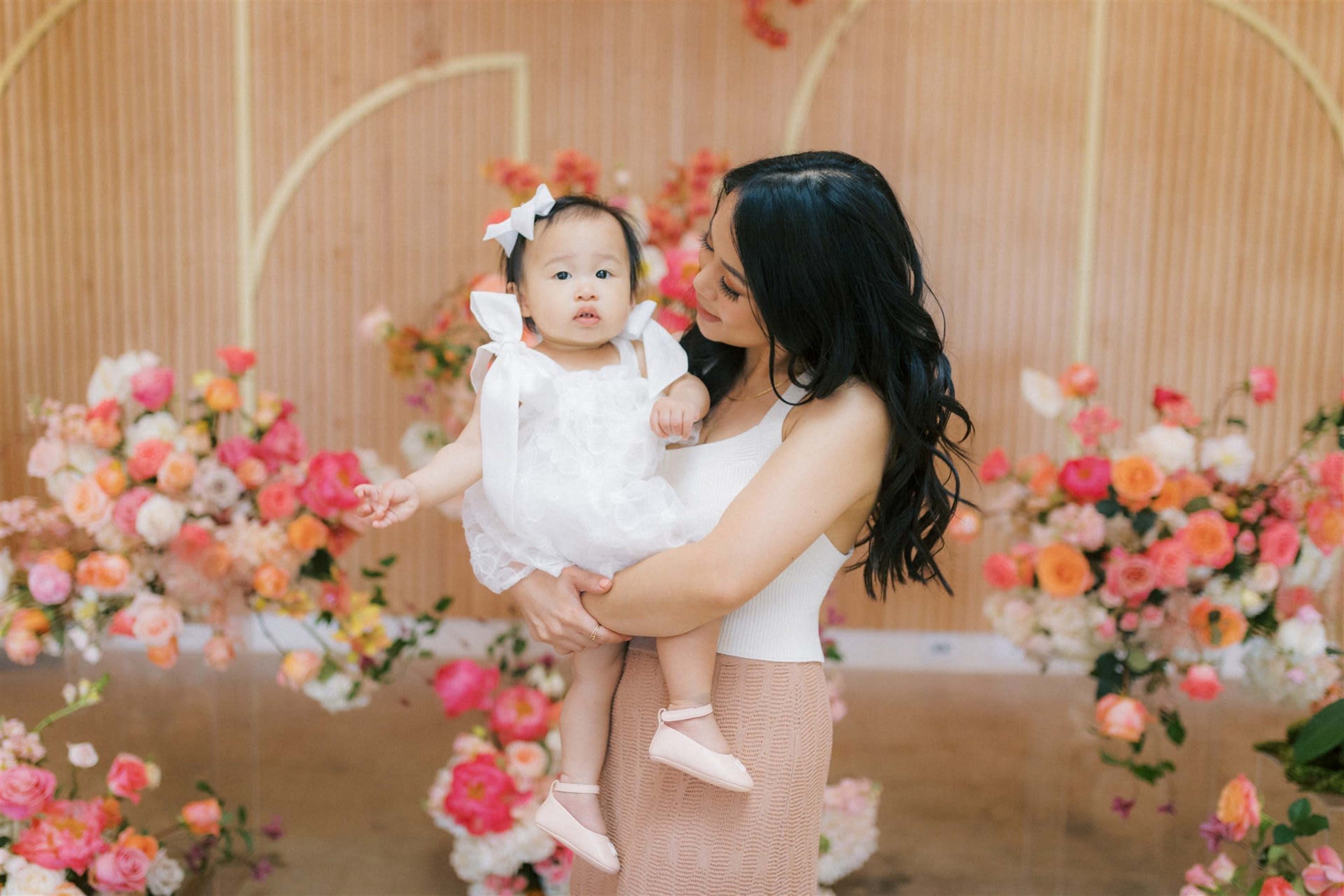 A woman holds a baby girl in a white dress surrounded by a colorful floral backdrop.