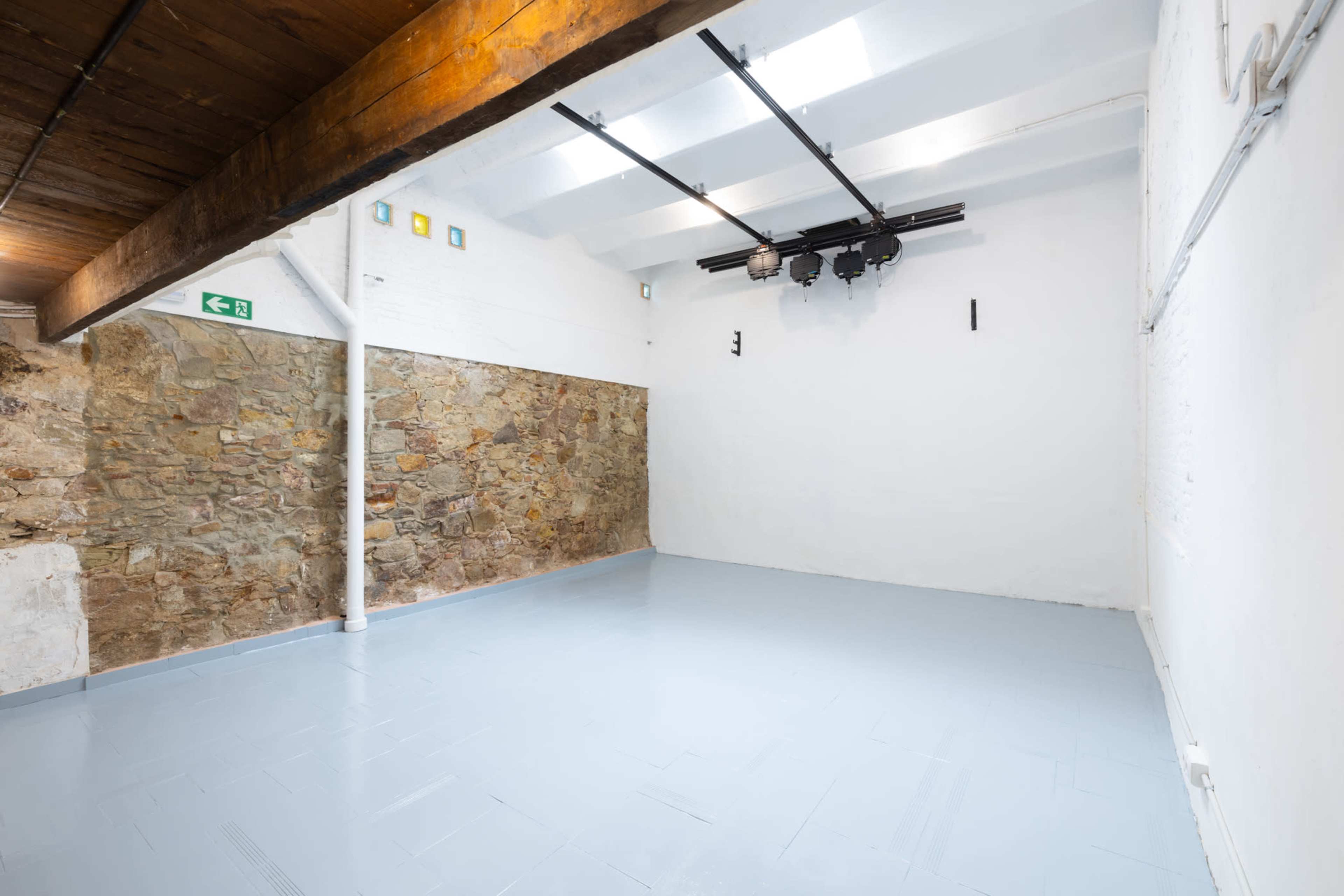 The image shows a spacious, empty room with a smooth blue floor, exposed wooden beams on the ceiling, and a stone wall on one side.