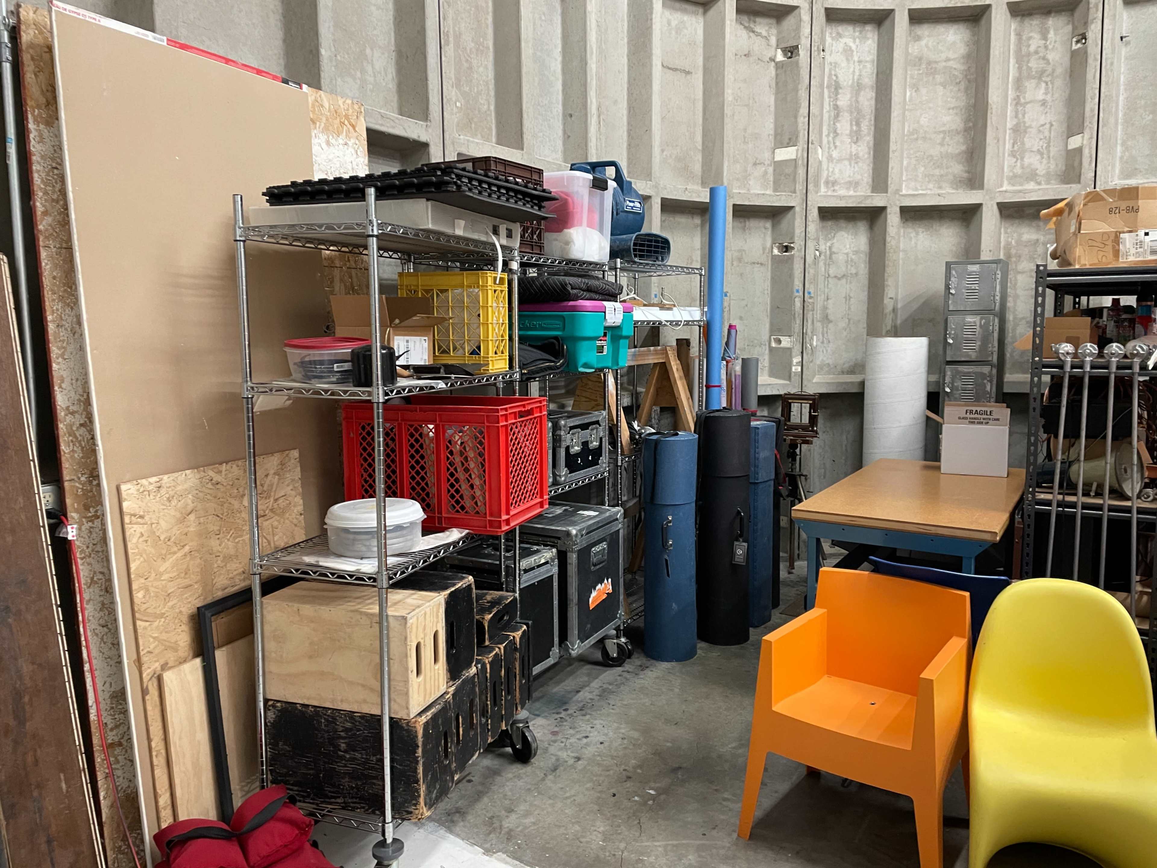 A cluttered storage area features shelves filled with various containers, tools, and equipment alongside stacked crates and a bright orange chair.
