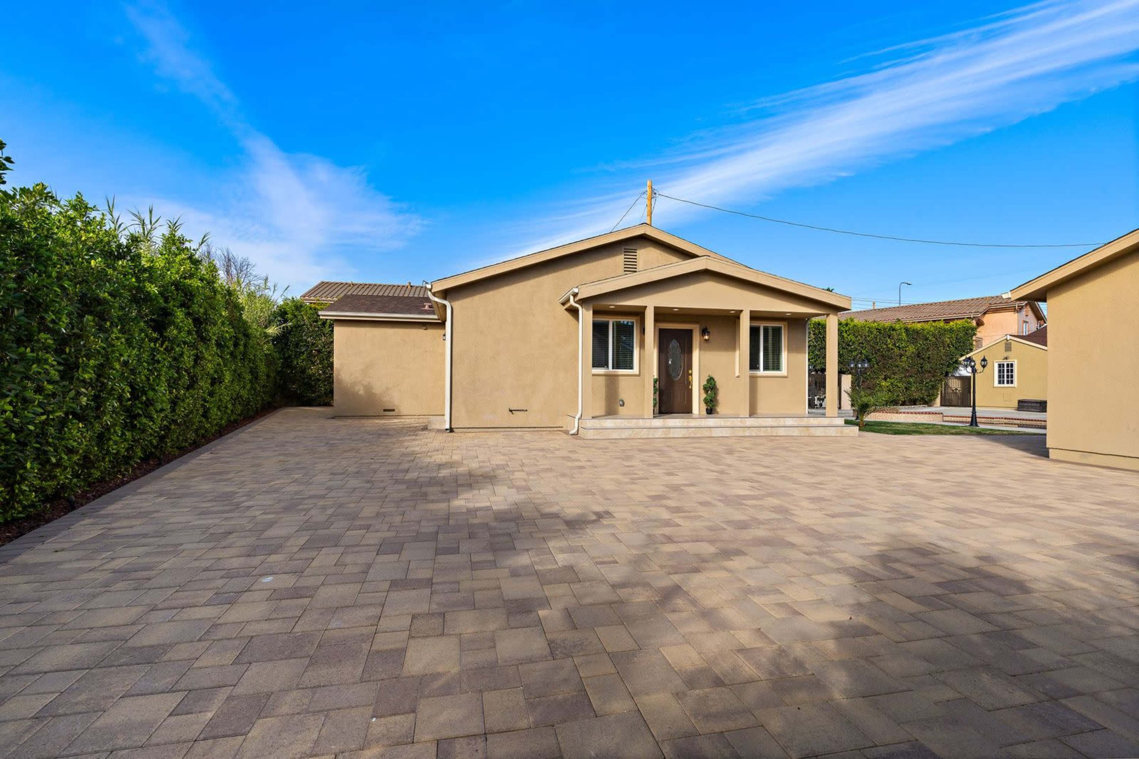 A single-story house with a light-colored exterior is situated on a paved driveway surrounded by low greenery under a blue sky.