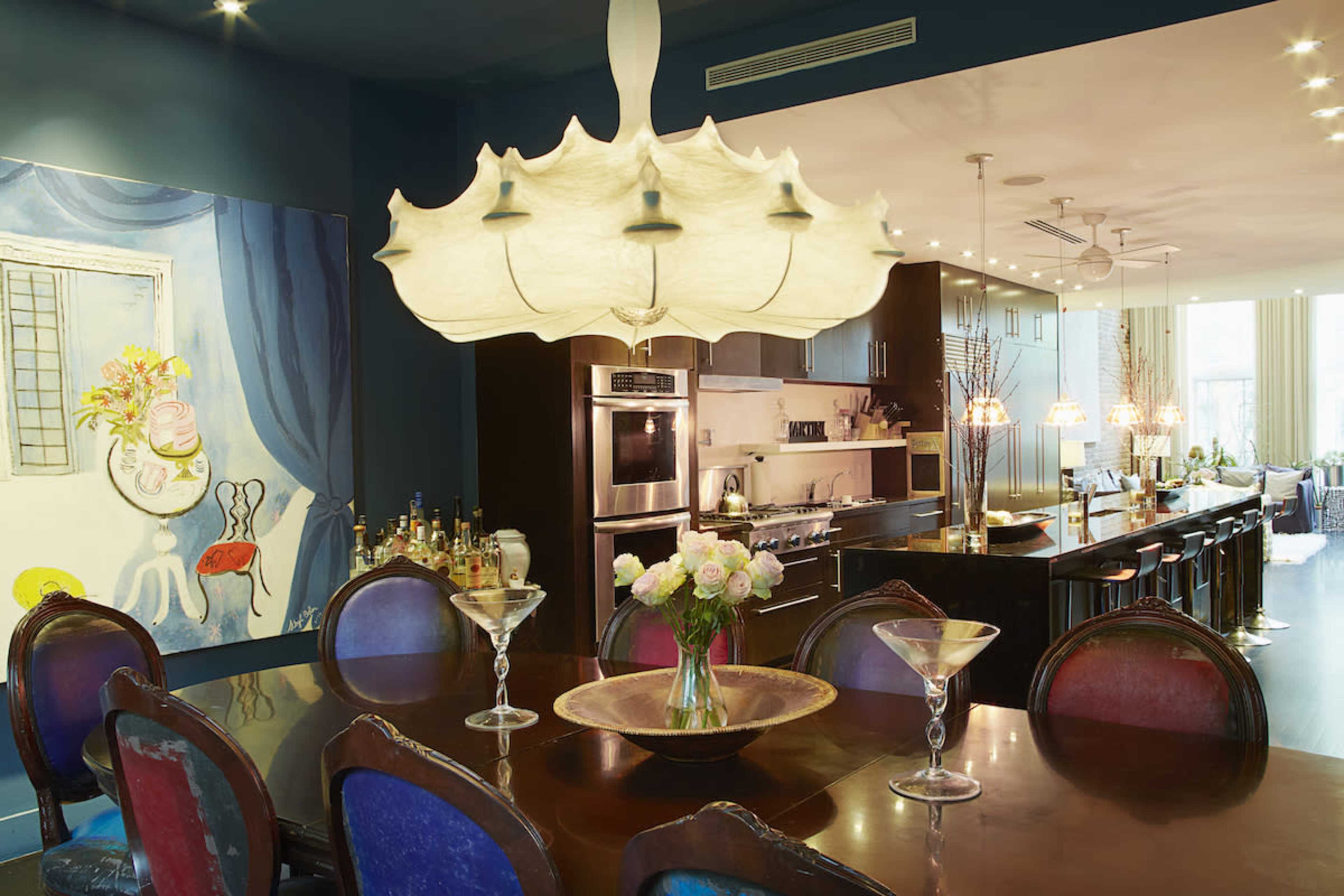 A modern kitchen and dining area featuring a large wooden table with colorful chairs, a decorative chandelier, and a vibrant painting on the wall.