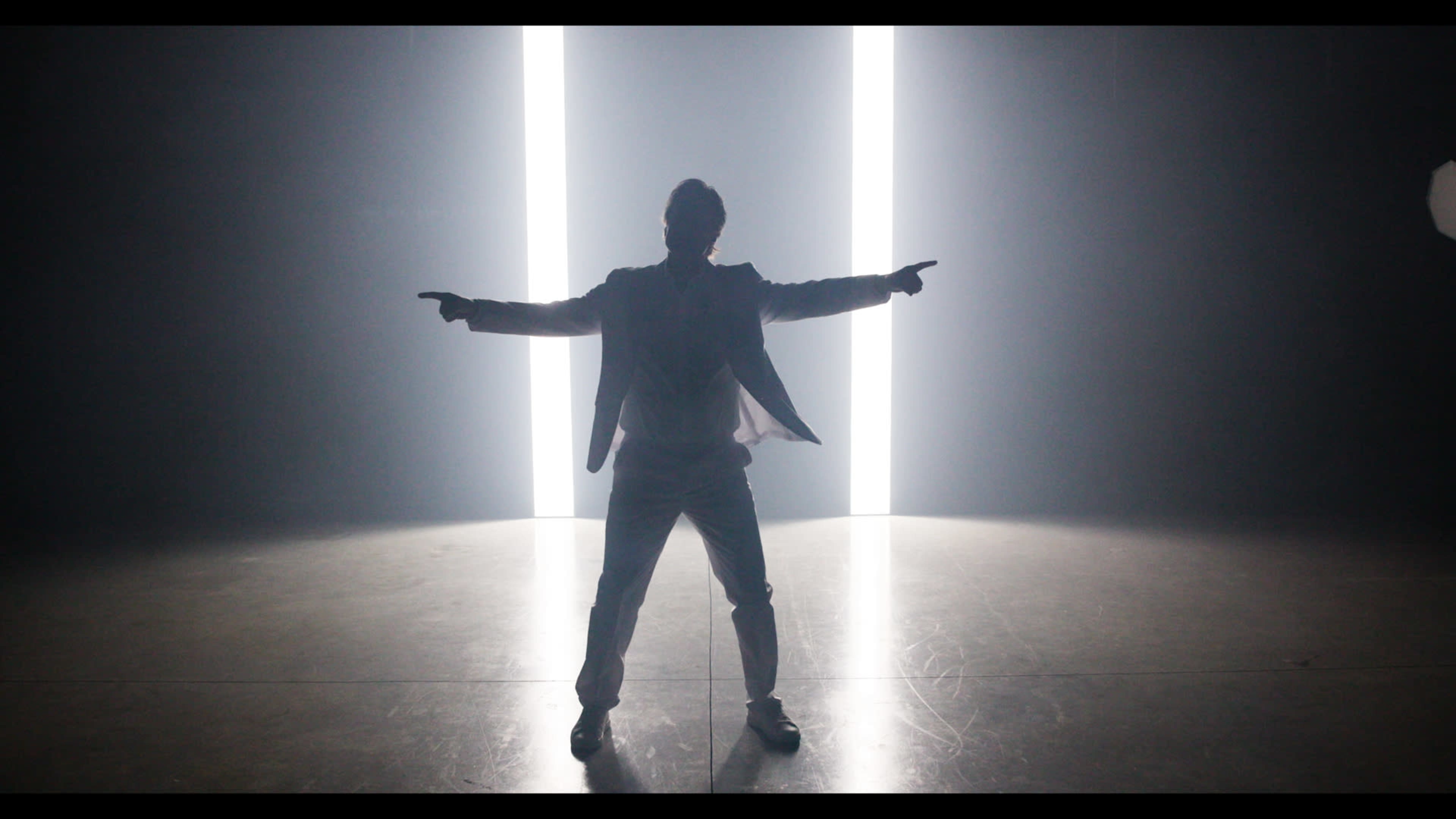 A silhouetted figure in a suit stands with outstretched arms between two bright vertical light beams on a dimly lit stage.