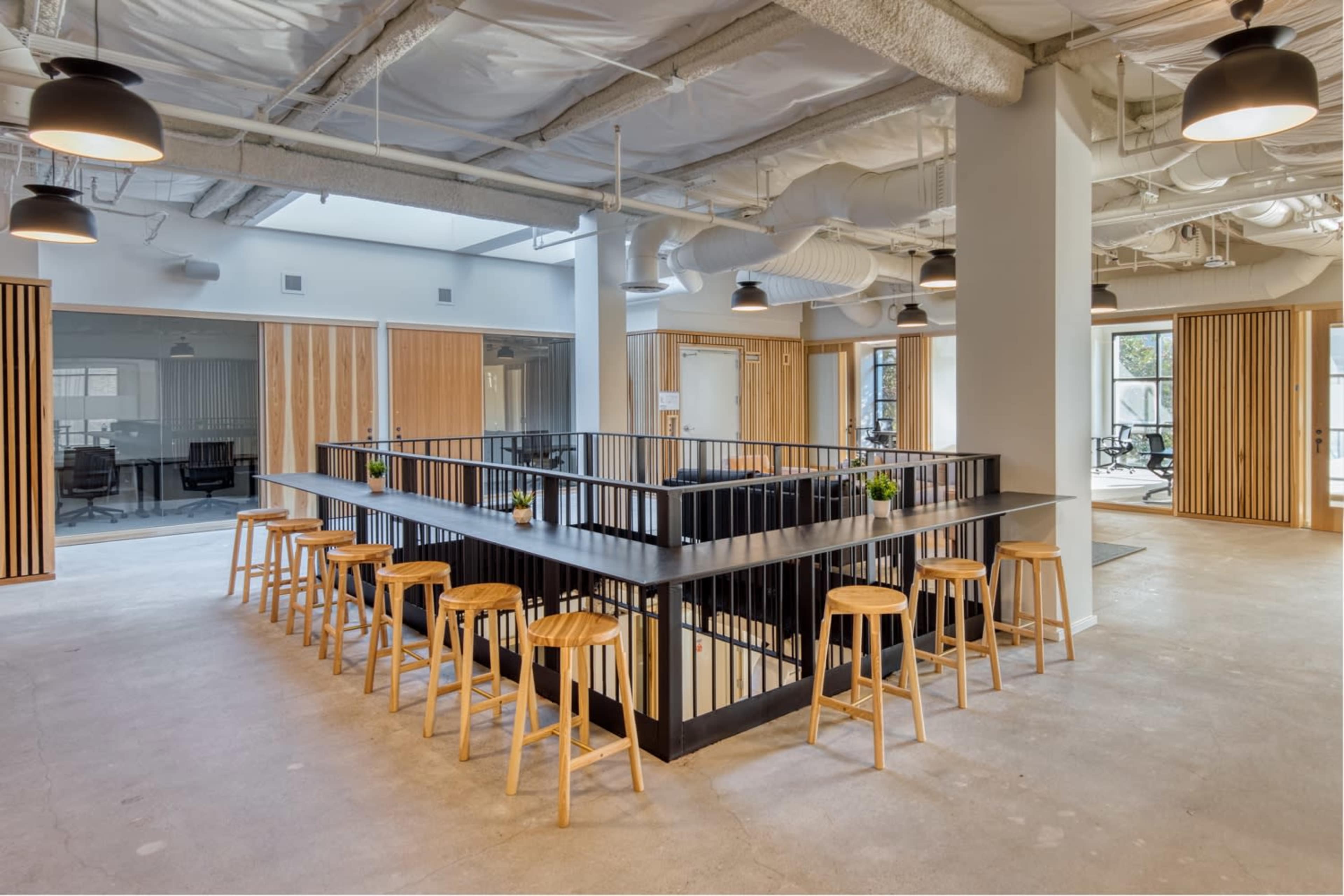 A modern office interior featuring a central bar-style table with wooden stools, surrounded by wooden paneling and large glass meeting rooms.