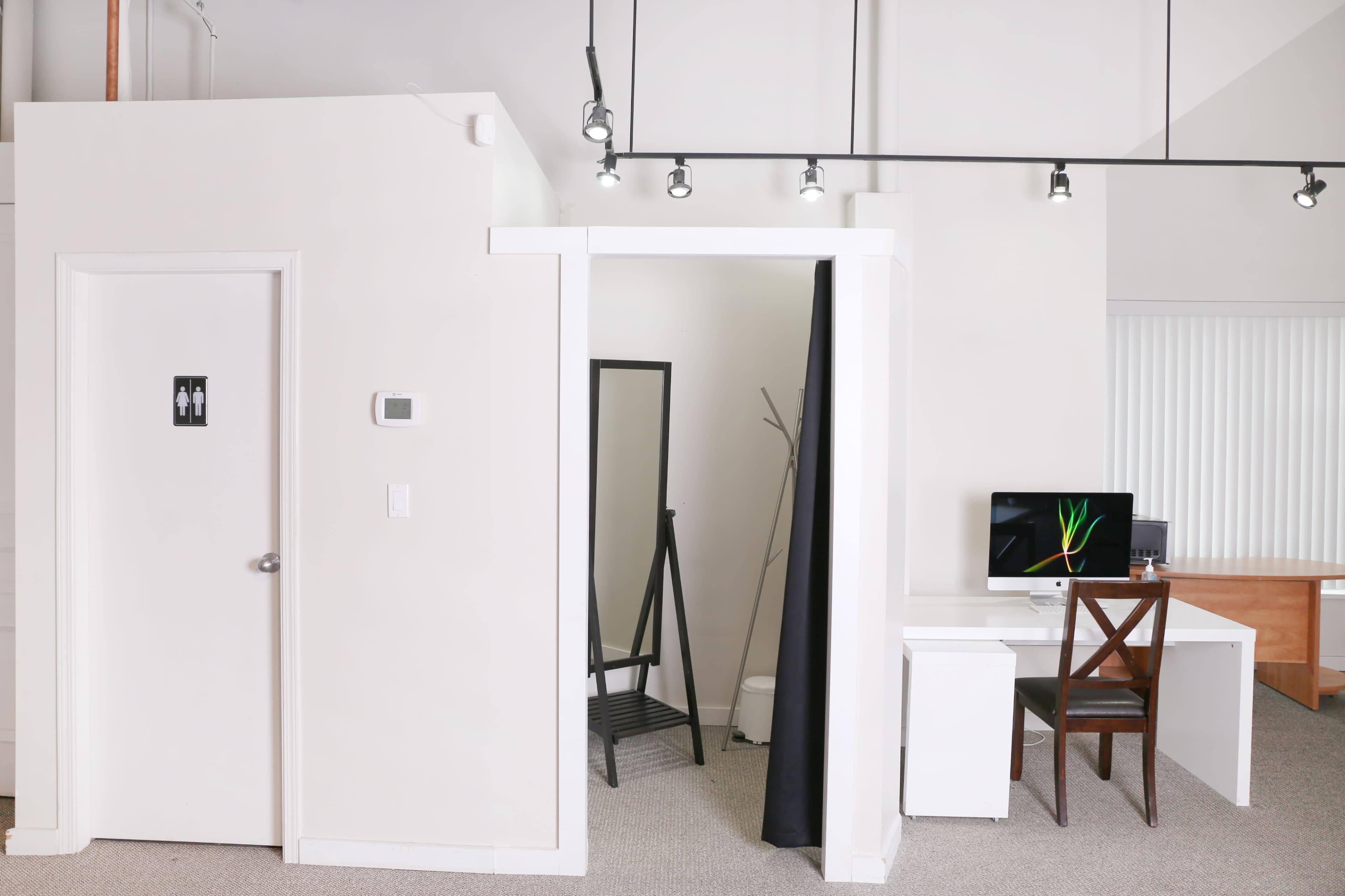 A bright, minimalistic room featuring a bathroom door, a standing mirror, a desk with a computer, and a chair.