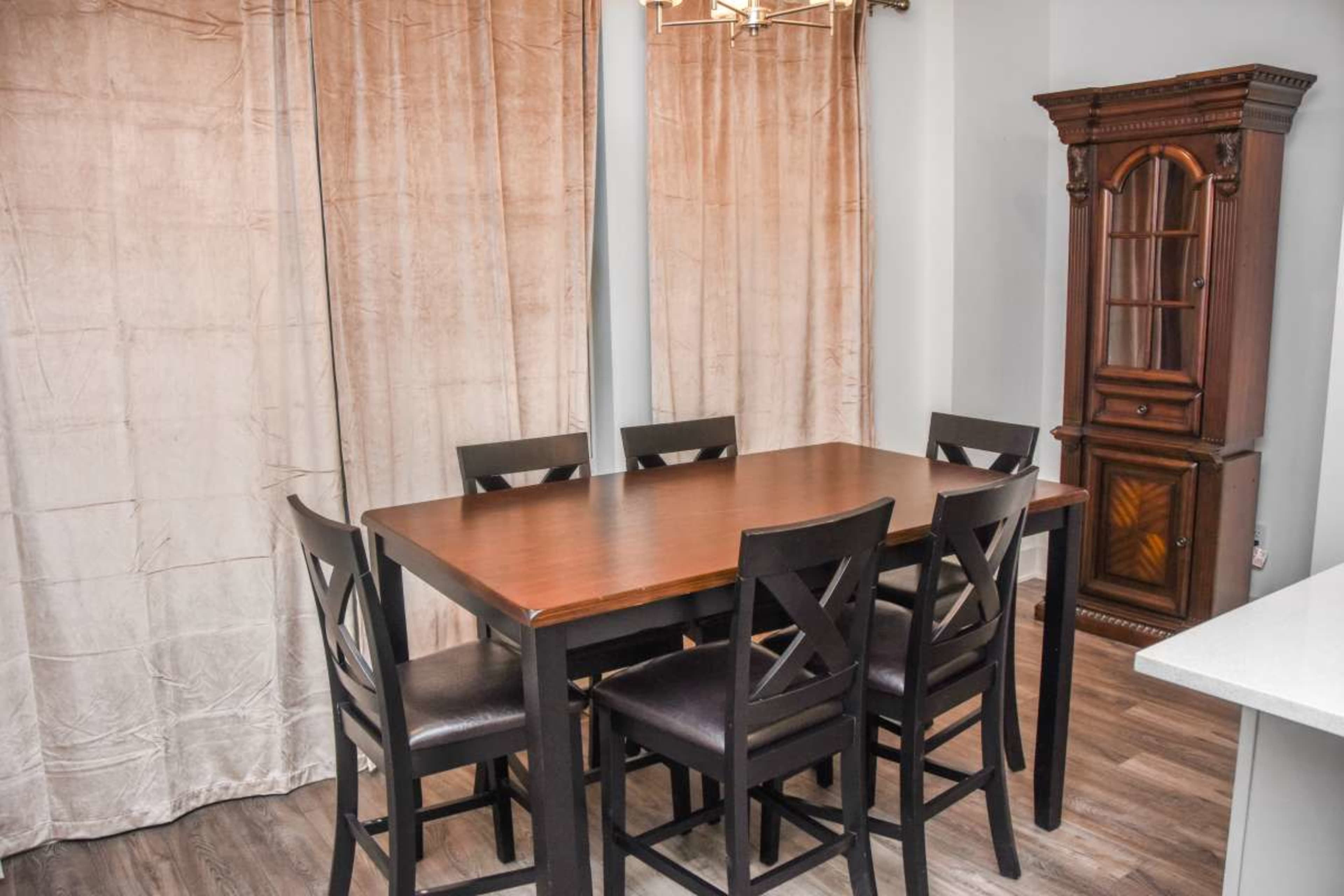 A wooden dining table surrounded by six black chairs is positioned in a room with beige curtains and a wooden display cabinet.