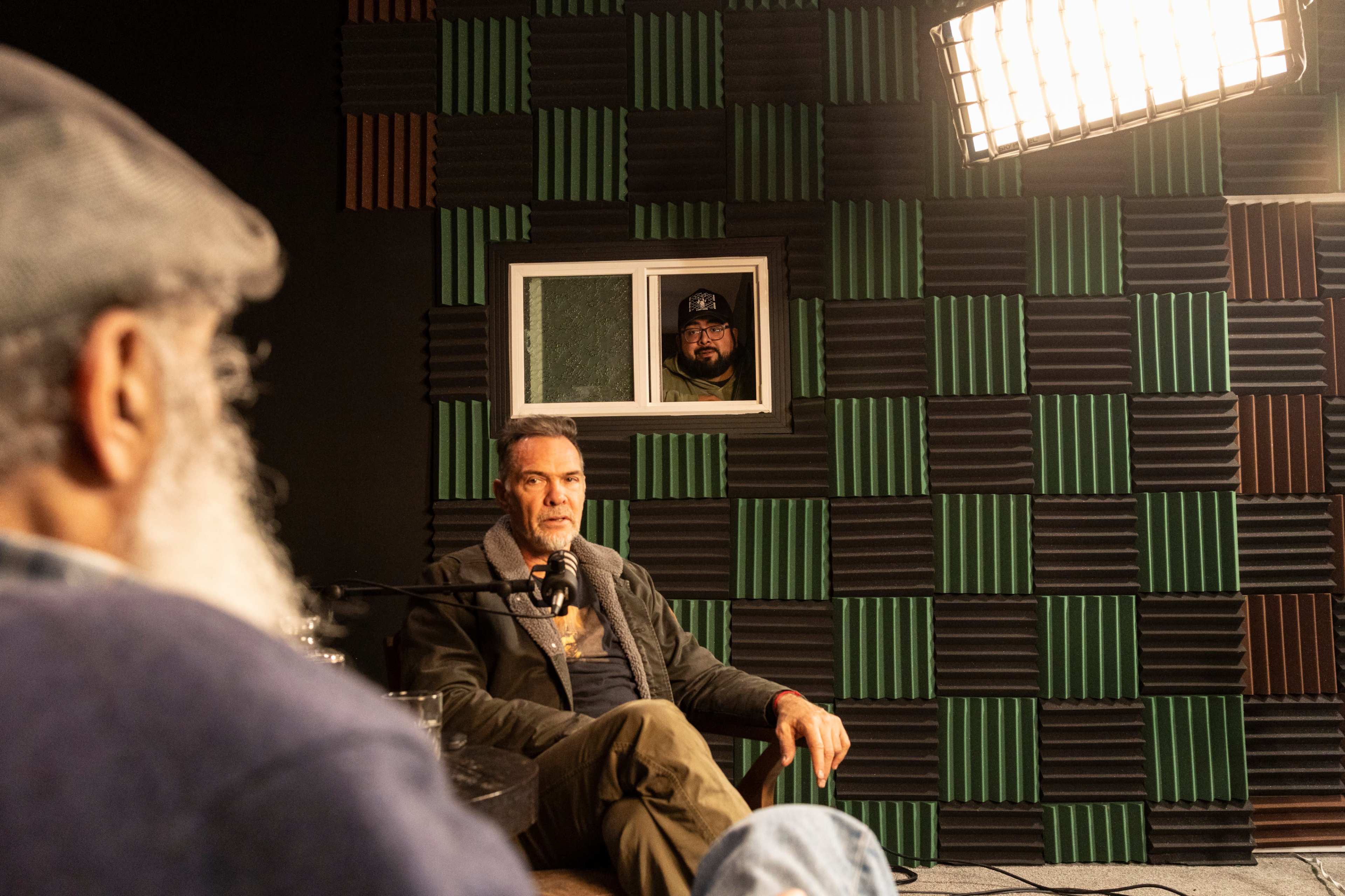 A podcast recording session takes place in a studio with soundproofed walls and a participant visible through a window in the background.