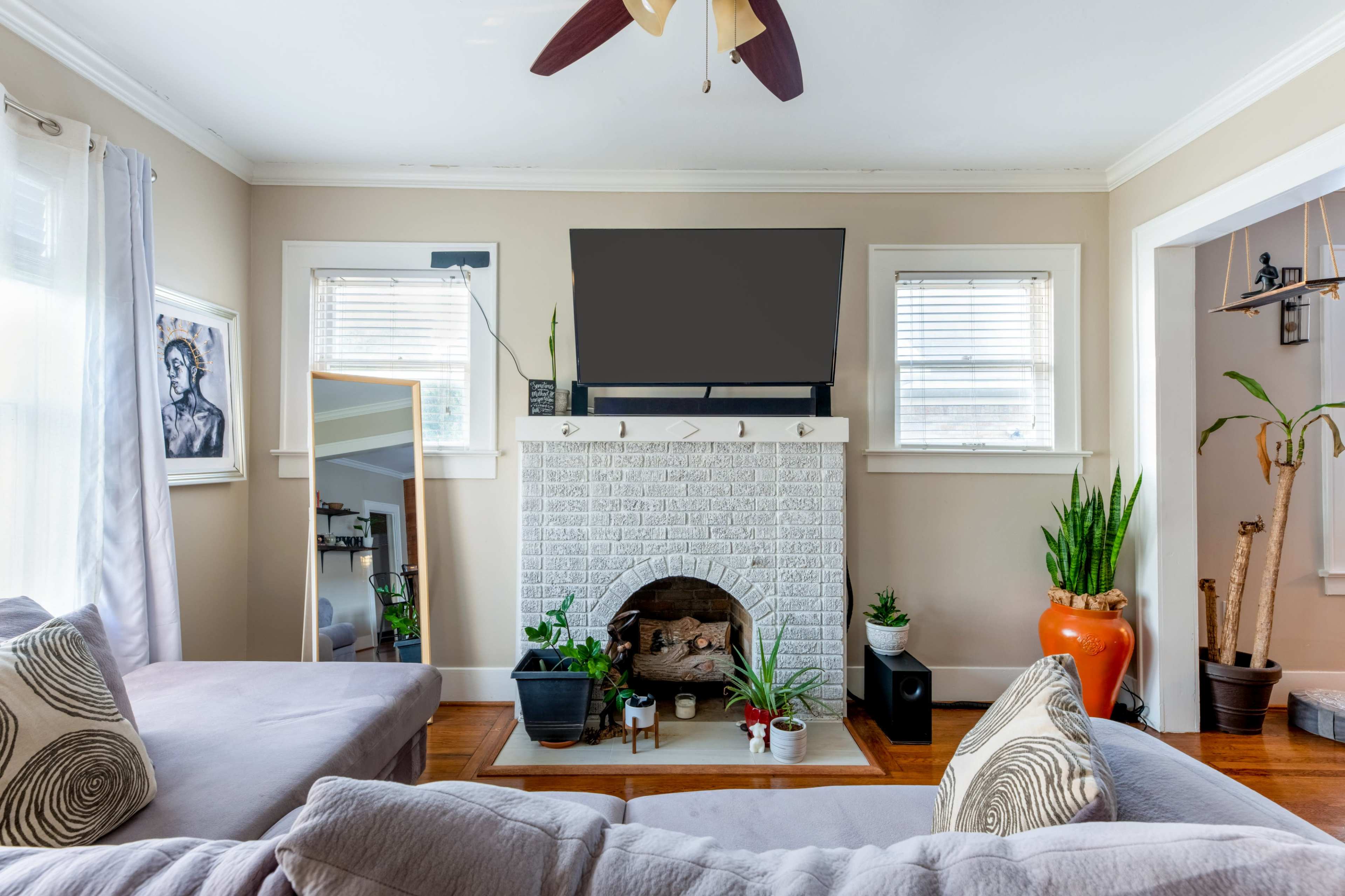 The living room features a white brick fireplace beneath a large television, with a sectional sofa and various indoor plants arranged around the space.