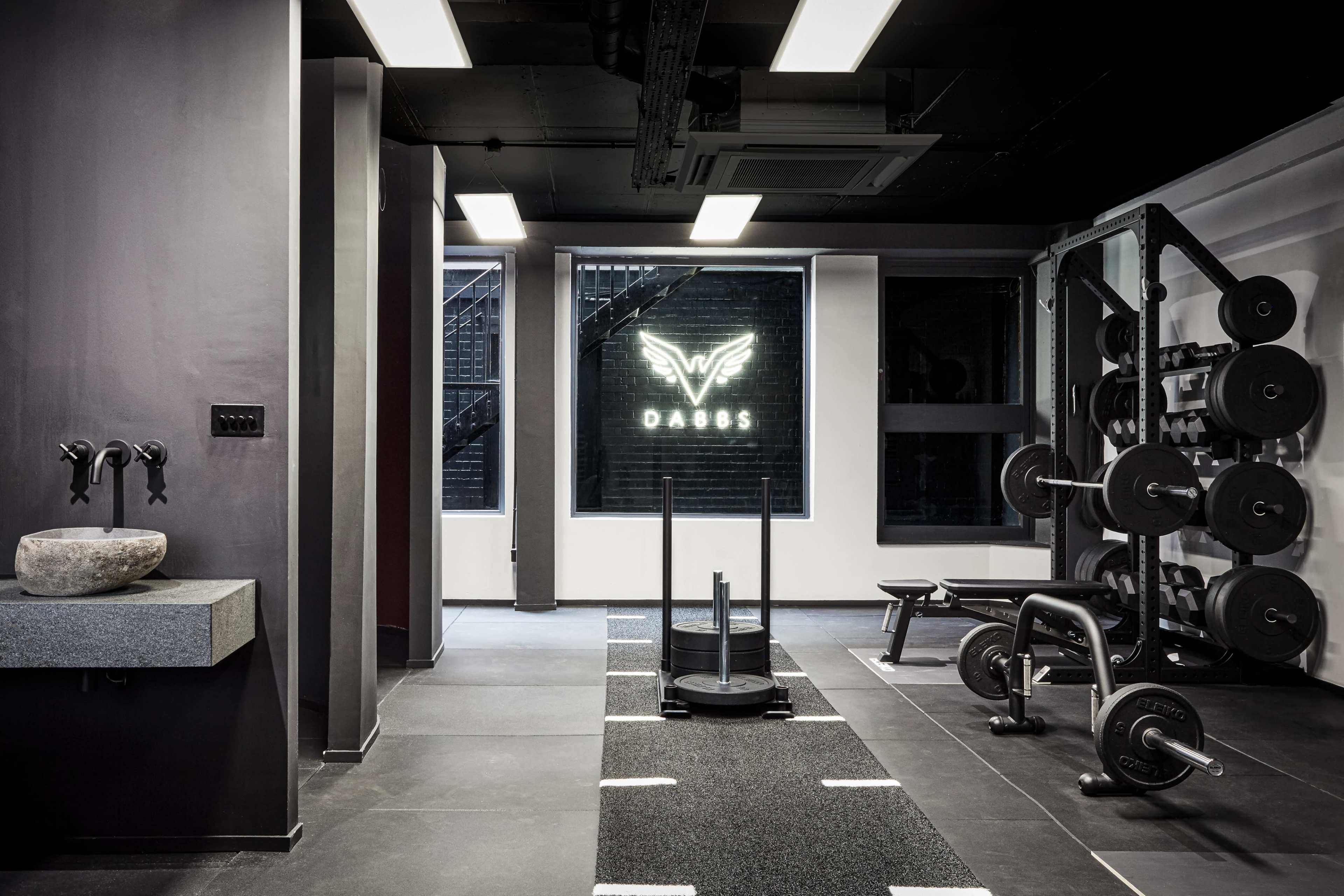 A modern gym interior with free weights, a sled, and a stone sink, featuring a glowing "DAOS" logo on the wall.