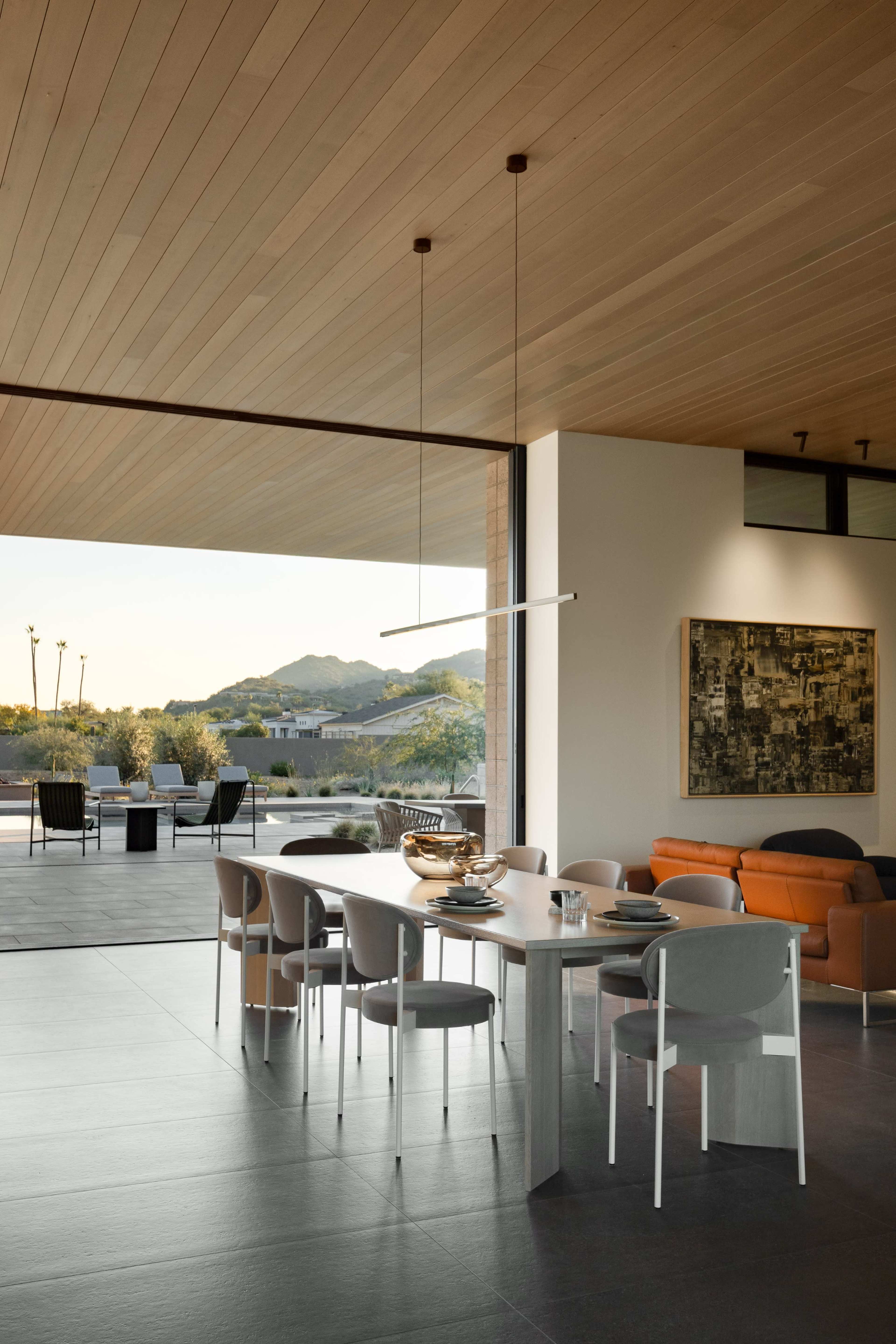 A modern dining area features a long table surrounded by minimalist chairs, with large glass doors leading to an outdoor space showcasing mountain views.