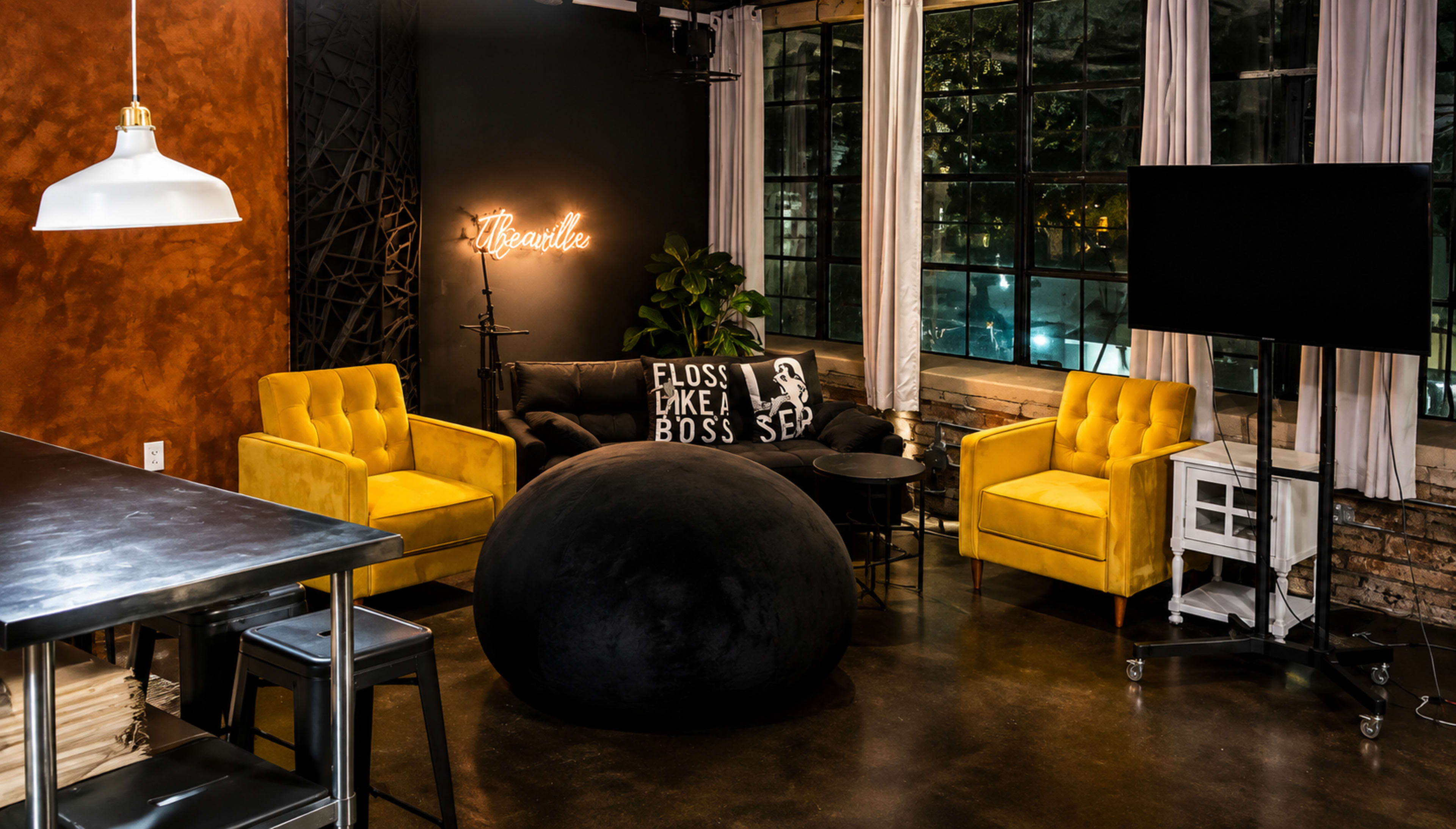 The image shows a modern lounge area featuring yellow chairs, a black sofa with decorative pillows, a large black bean bag, and a window with a view of the outdoors.