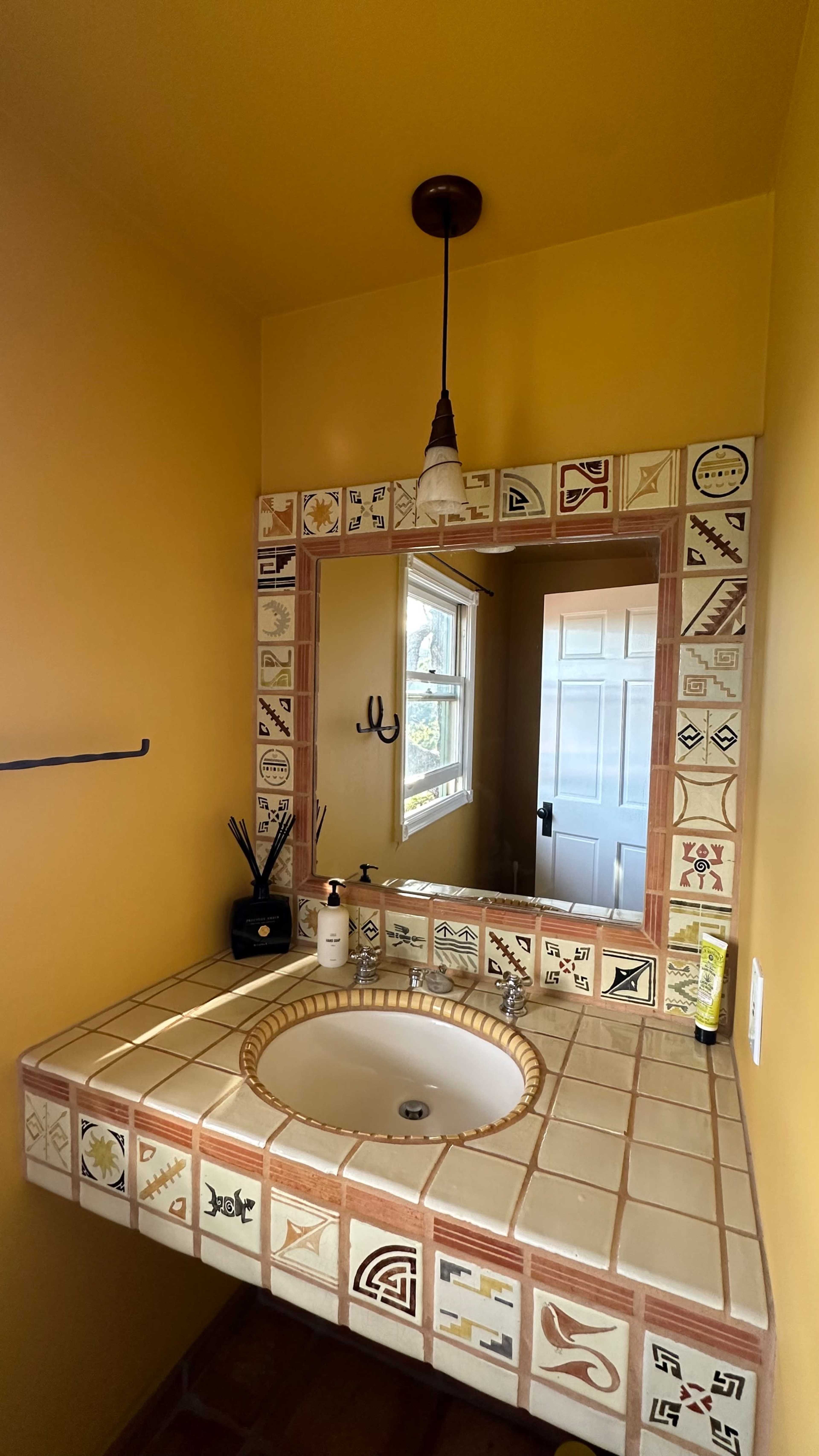A bathroom with a yellow wall, a tile sink surrounded by decorative tiles, and a mirror reflecting a door.