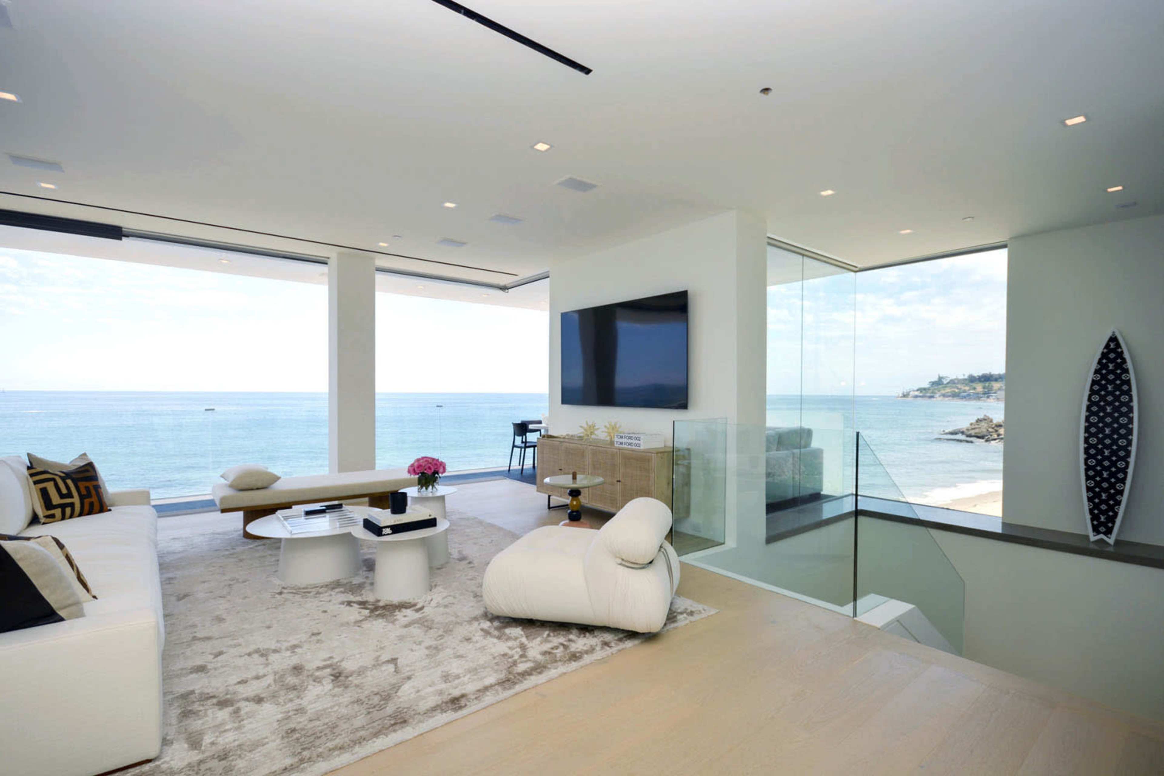 A modern living room features large windows overlooking the ocean, with minimalist furniture and a neutral color palette.