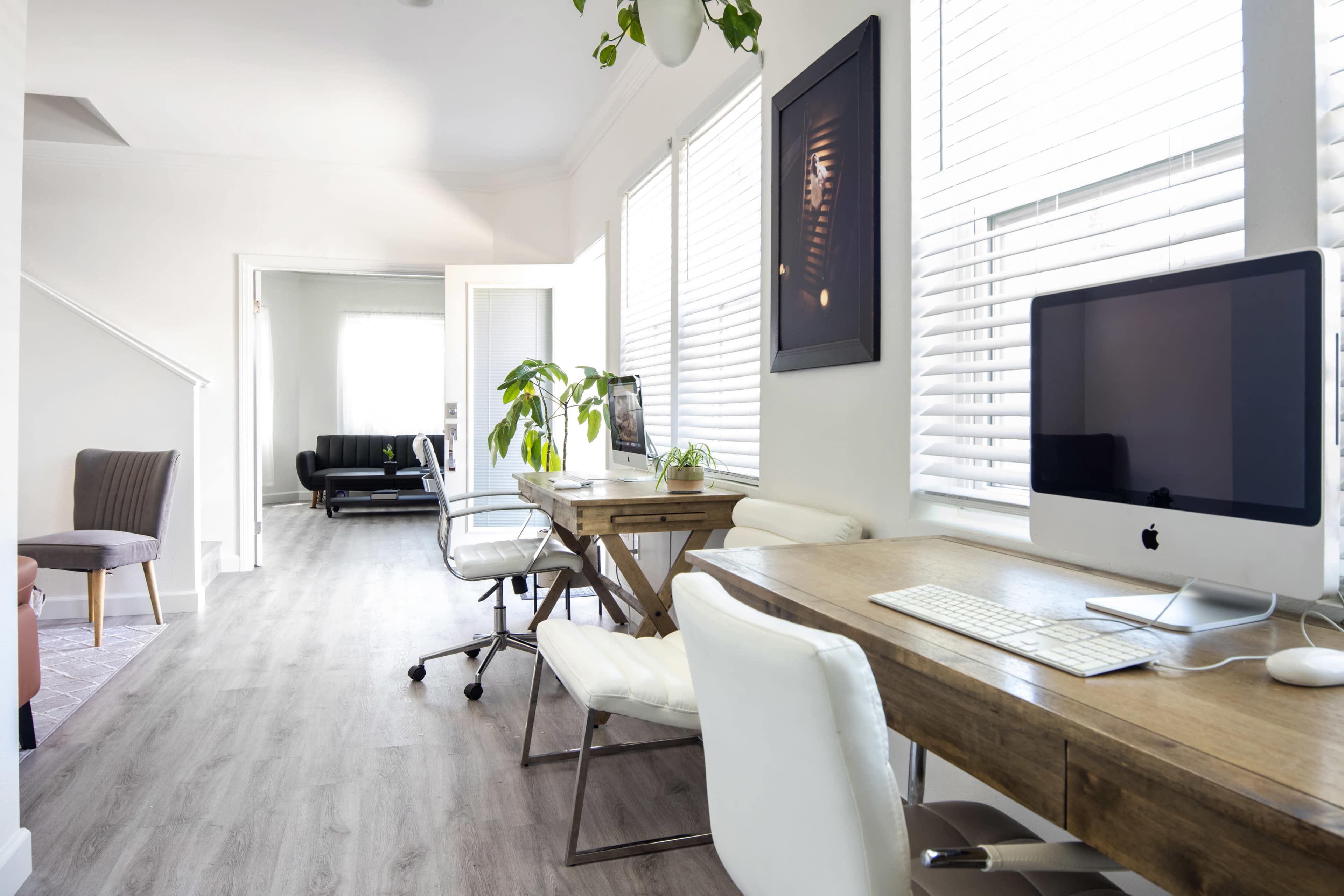 The image depicts a well-lit home office featuring two wooden desks with white chairs, an iMac computer, and a plant, leading to a sitting area with a black couch in the background.