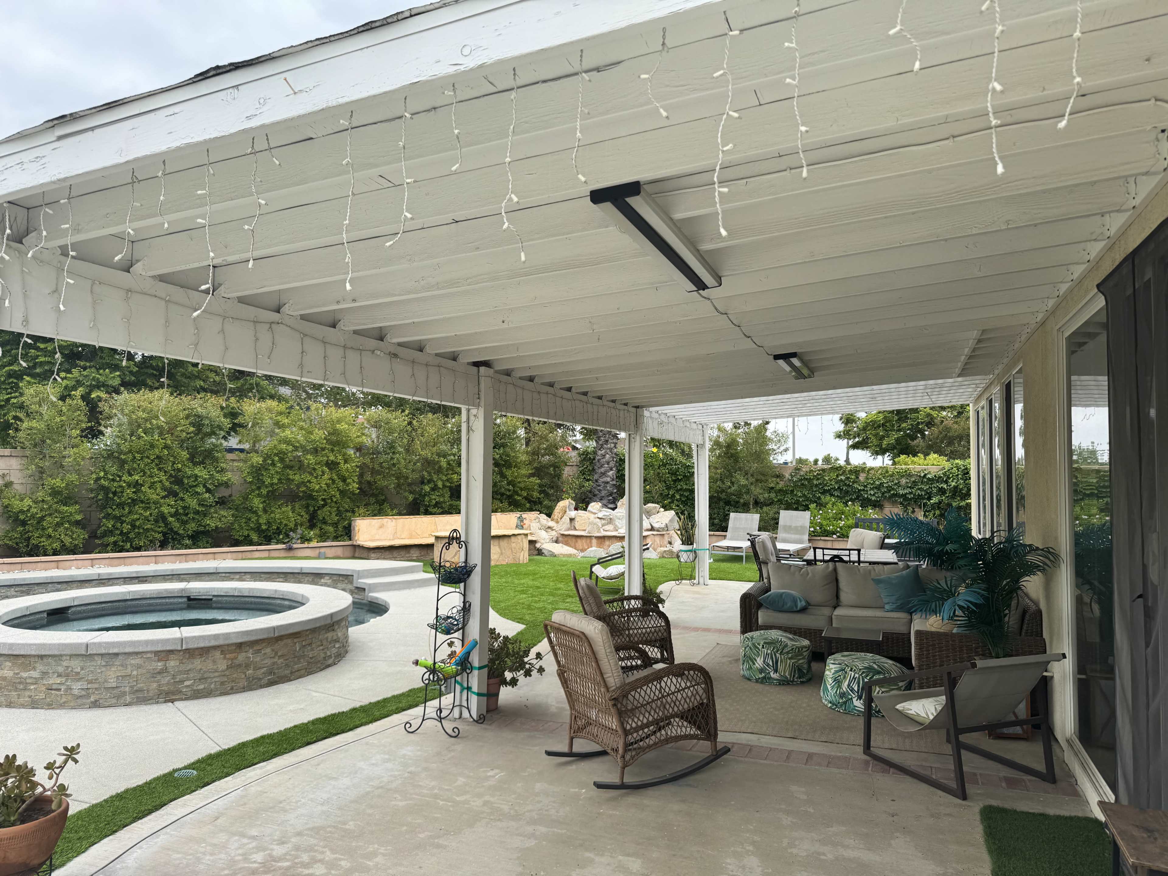 The image shows a covered patio area with seating, decorative plants, and a hot tub in the background.