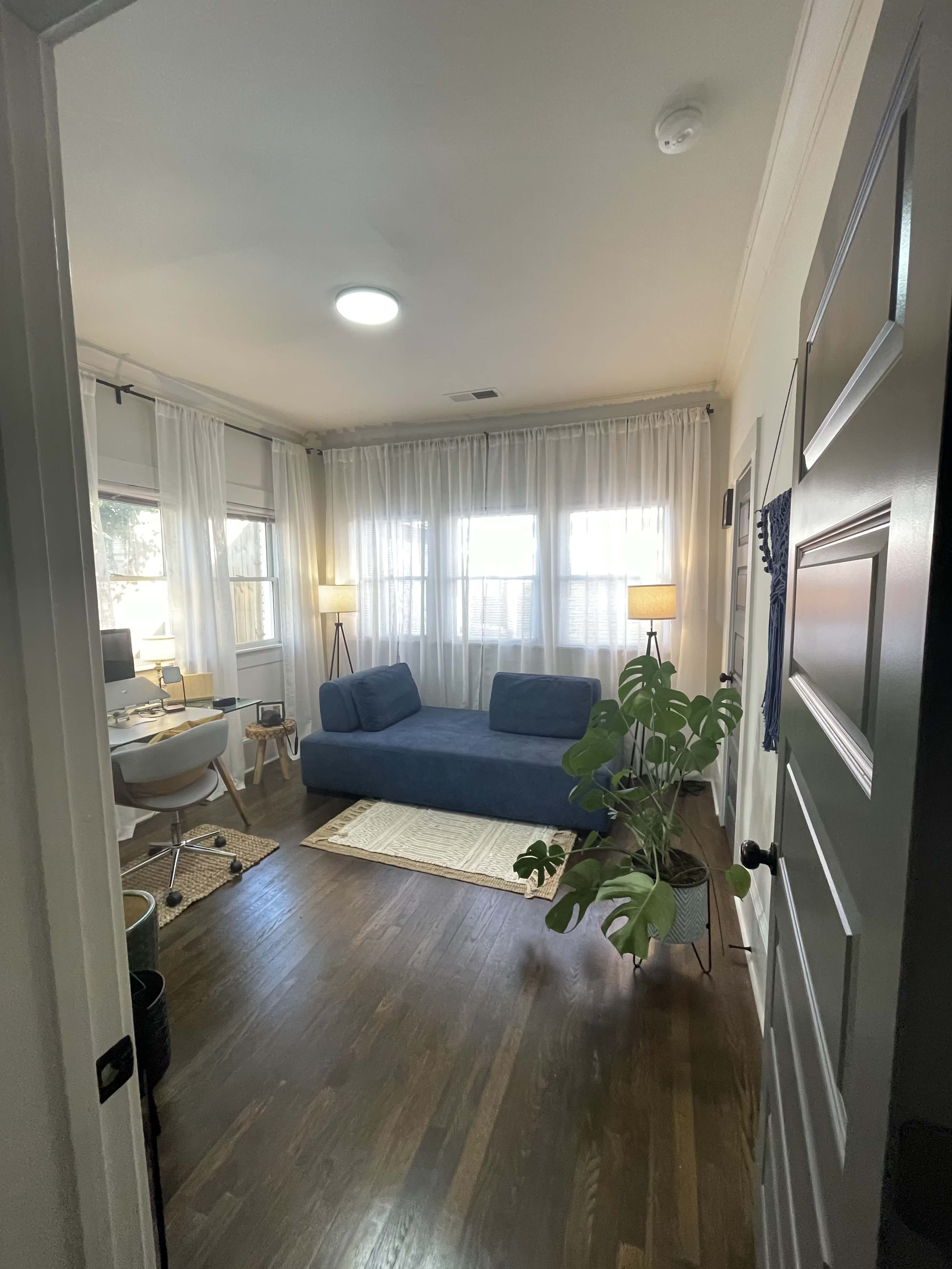 The image shows a brightly lit room with a blue couch, a potted plant, a desk with a computer, and two lamps, all arranged around a woven rug on a hardwood floor.