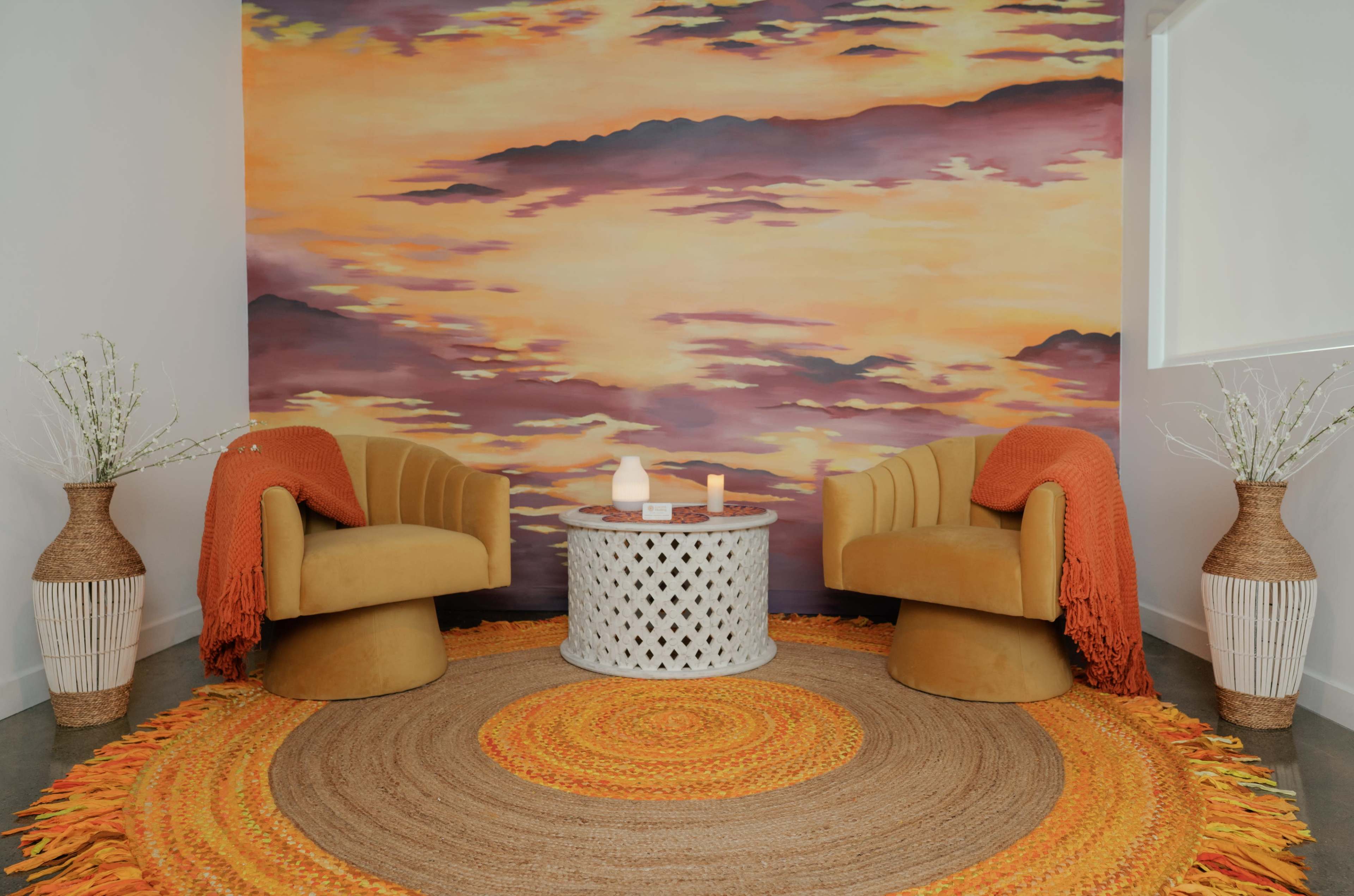 The image shows a cozy seating area with two upholstered chairs, a circular table in the center, and a large, colorful mural depicting a sunset.