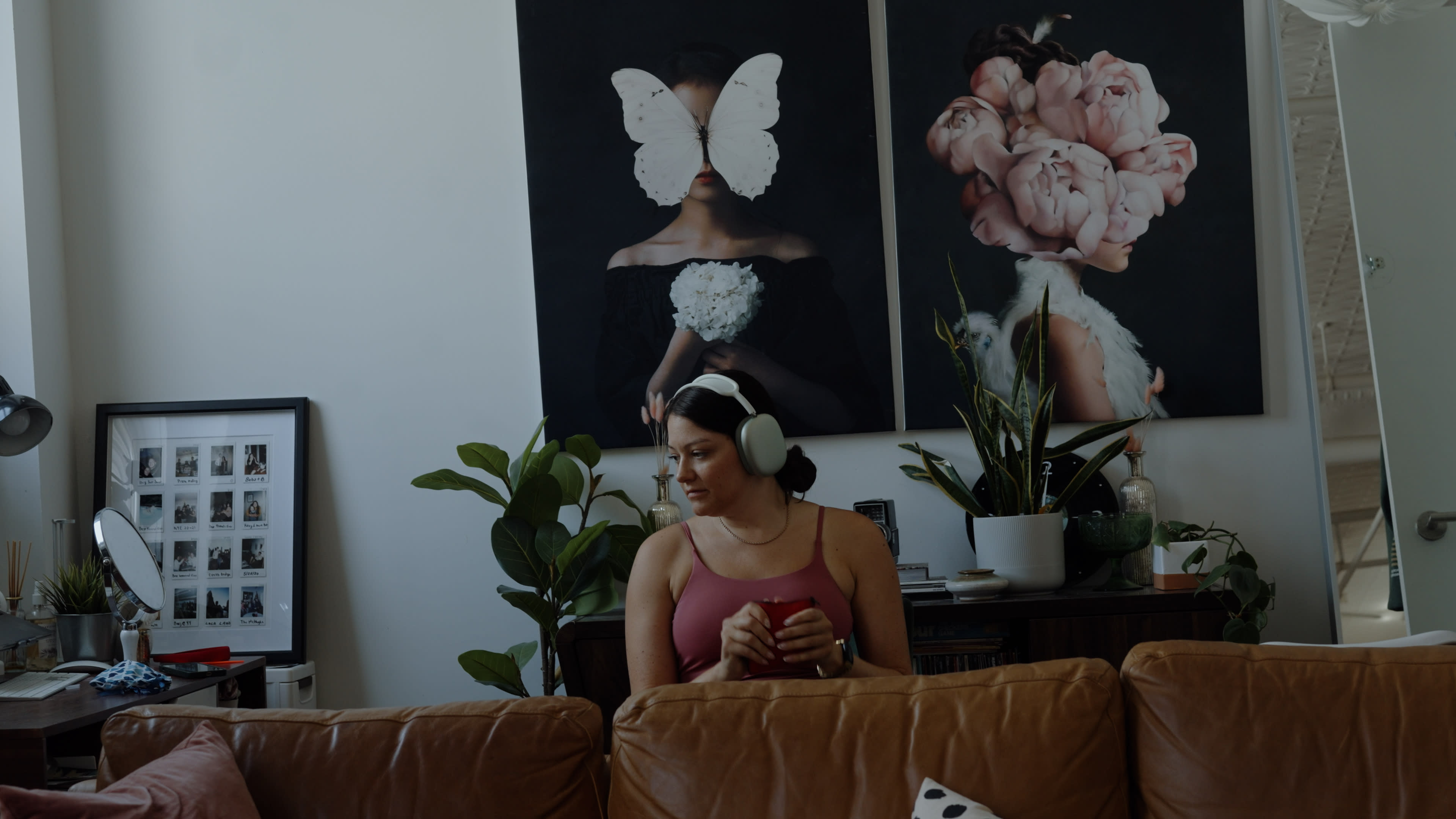 A woman in a pink tank top sits on a brown couch, wearing headphones and holding a mug, with large artworks of a woman with a butterfly and flowers on the wall behind her.