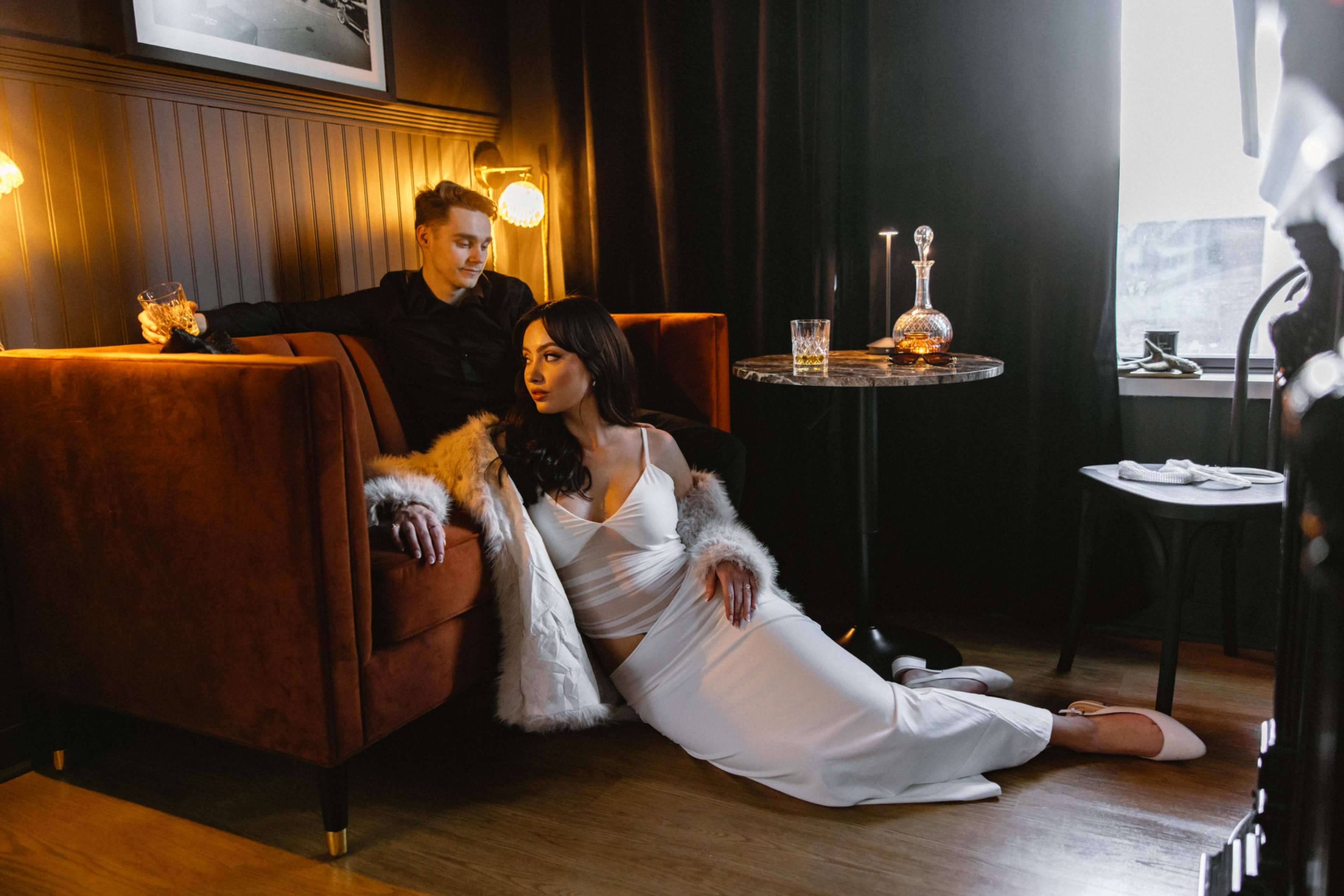 A man sits on a couch while a woman in a white dress lounges beside him, both in a dimly lit room with stylish decor.