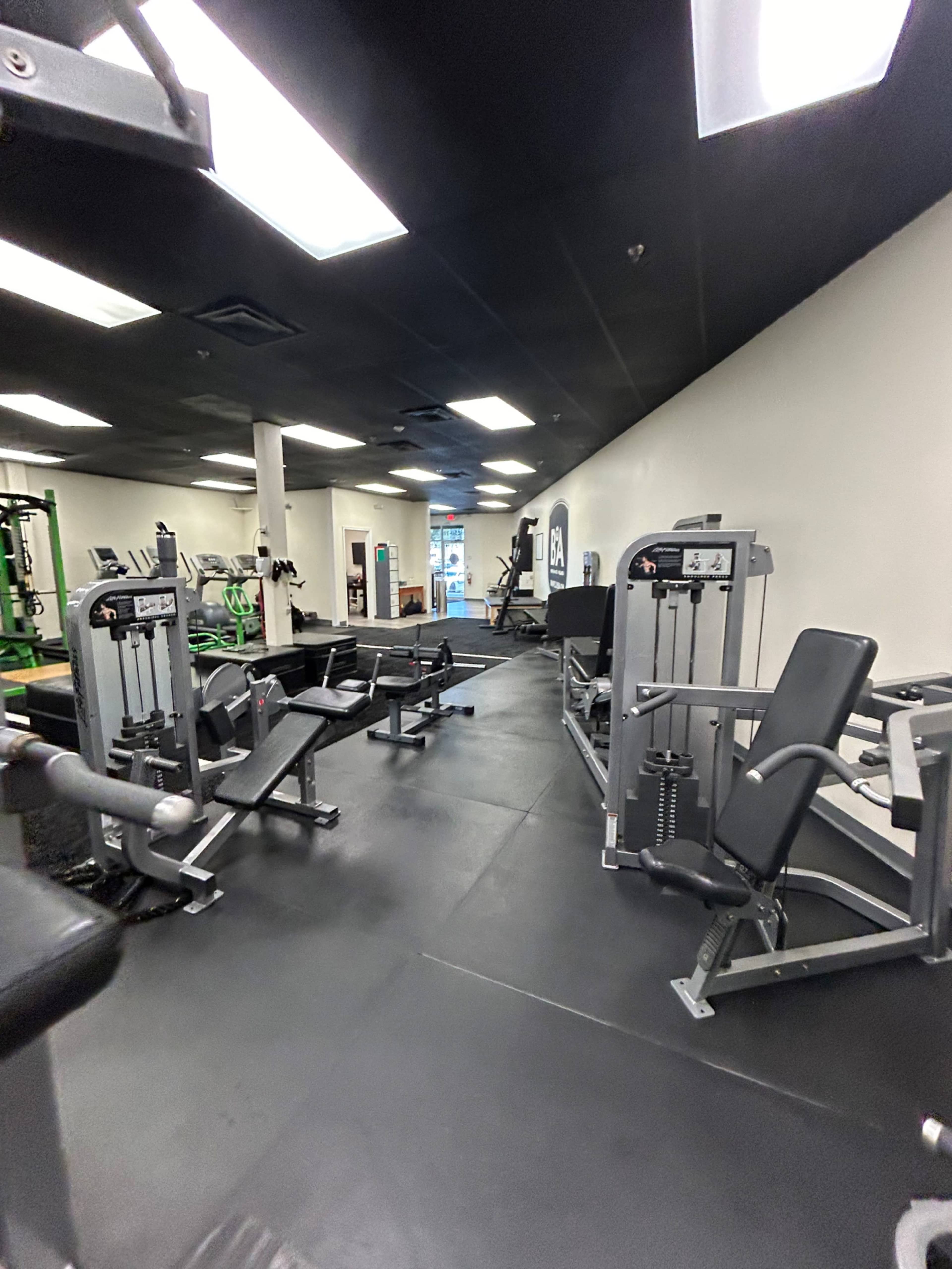 A gym interior with various weight training machines arranged on a black rubber floor under bright overhead lights.