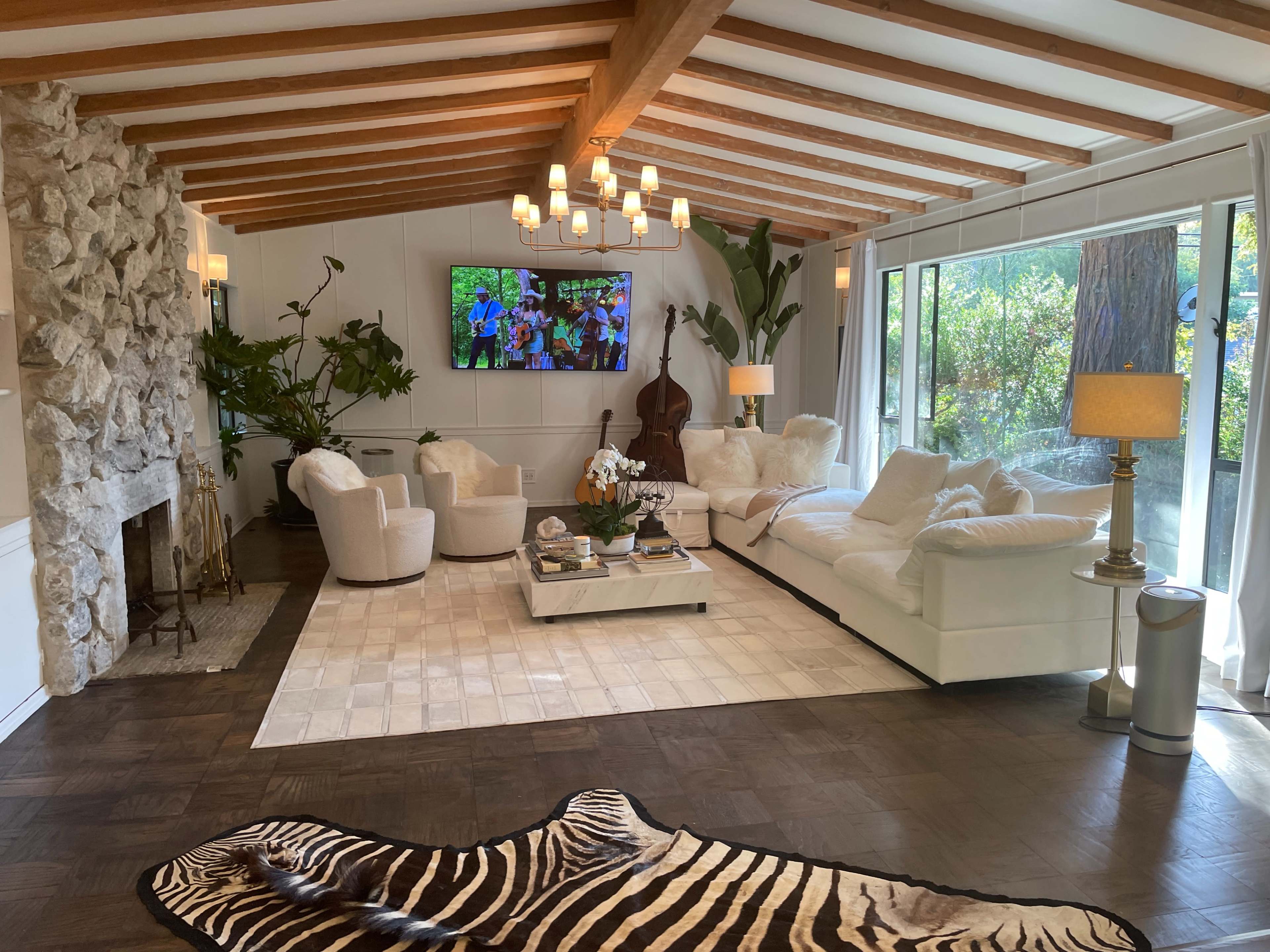 The image shows a modern living room with a stone fireplace, large windows, white furniture, and a patterned area rug.