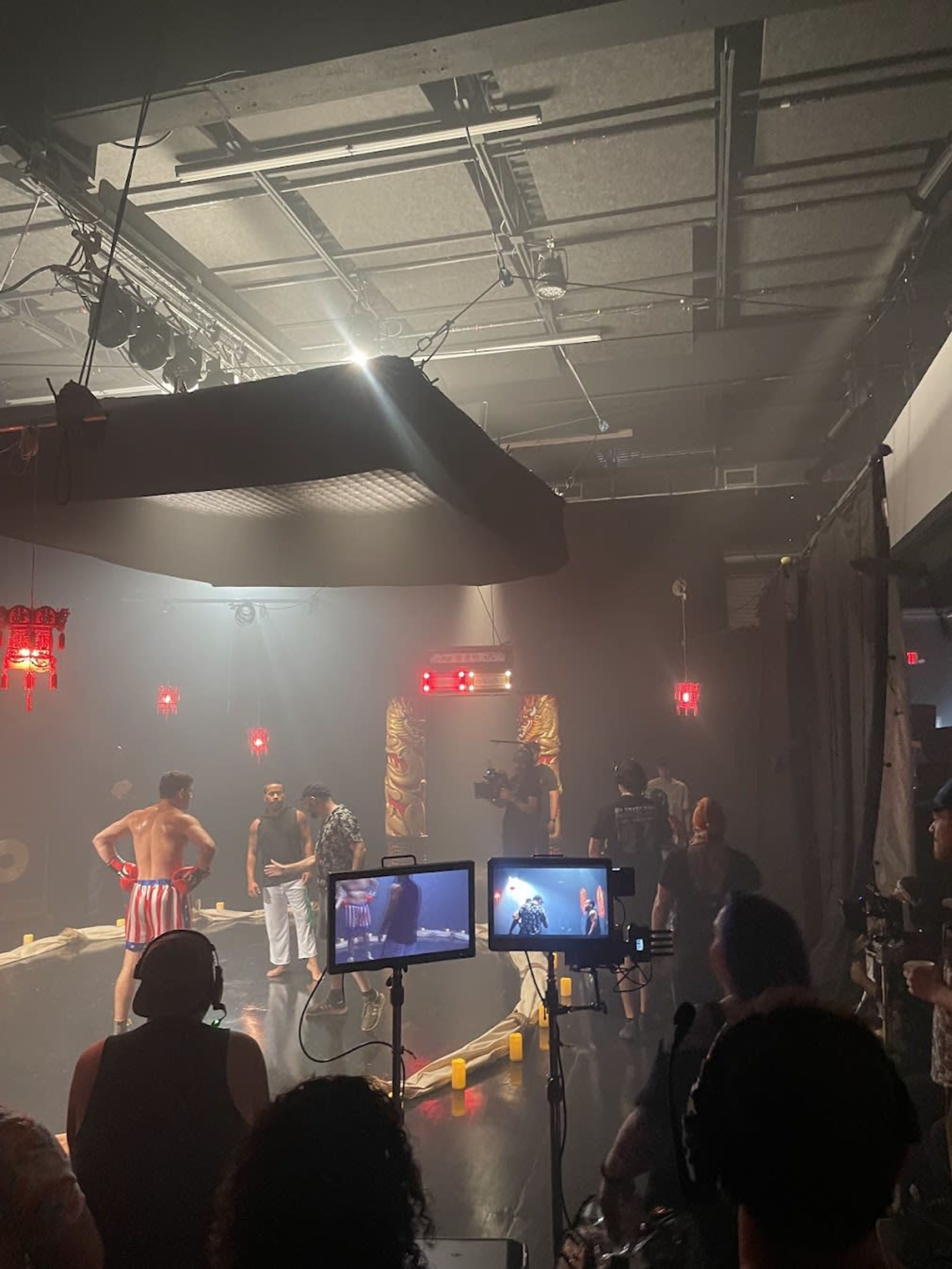 The image shows a film set with several crew members operating cameras while actors perform in front of a backdrop illuminated by red lights.