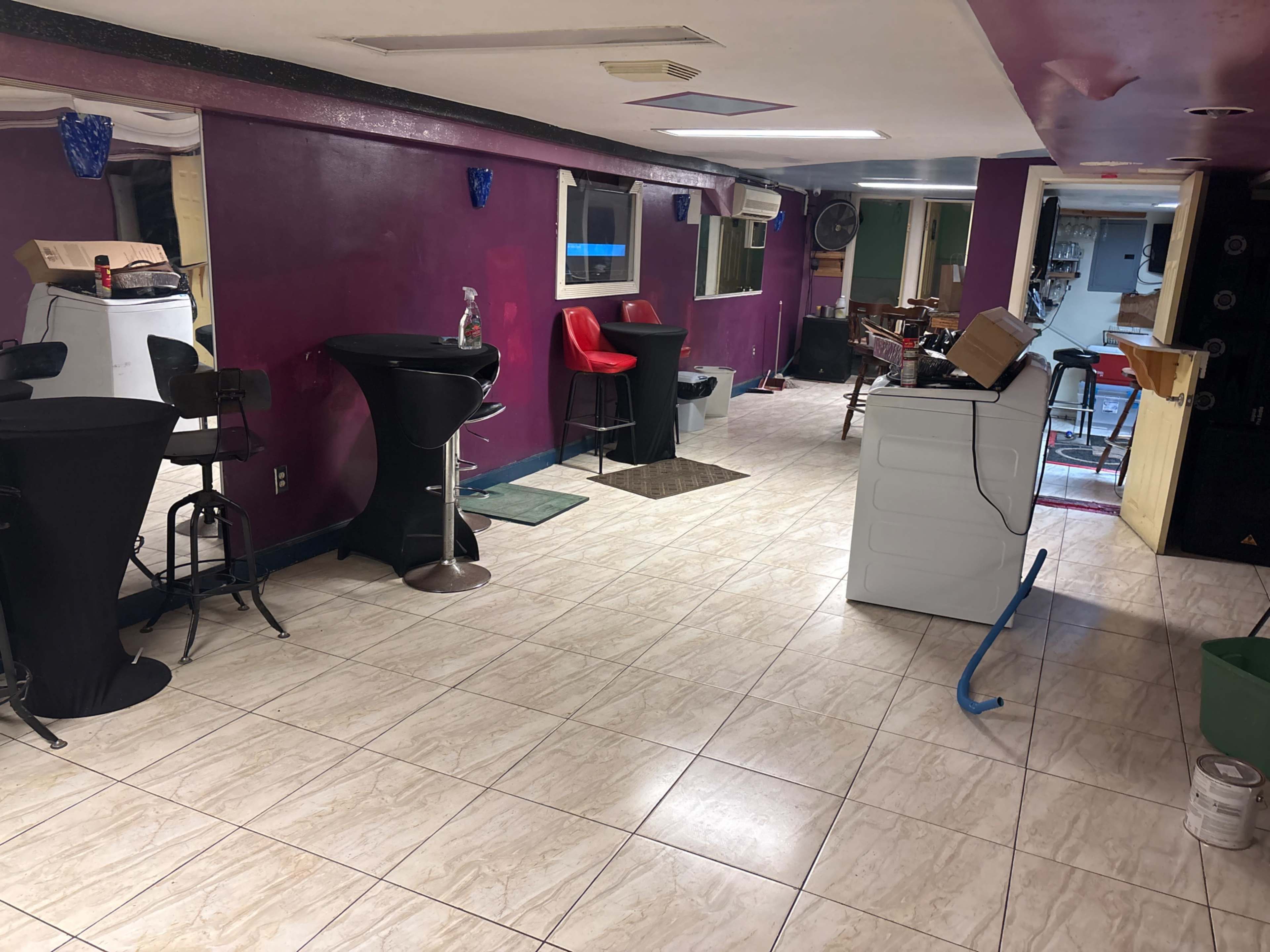 The image shows a large room with tiled flooring, featuring bar-style seating, a white washing machine, and colorful walls, with various furniture and equipment arranged throughout.