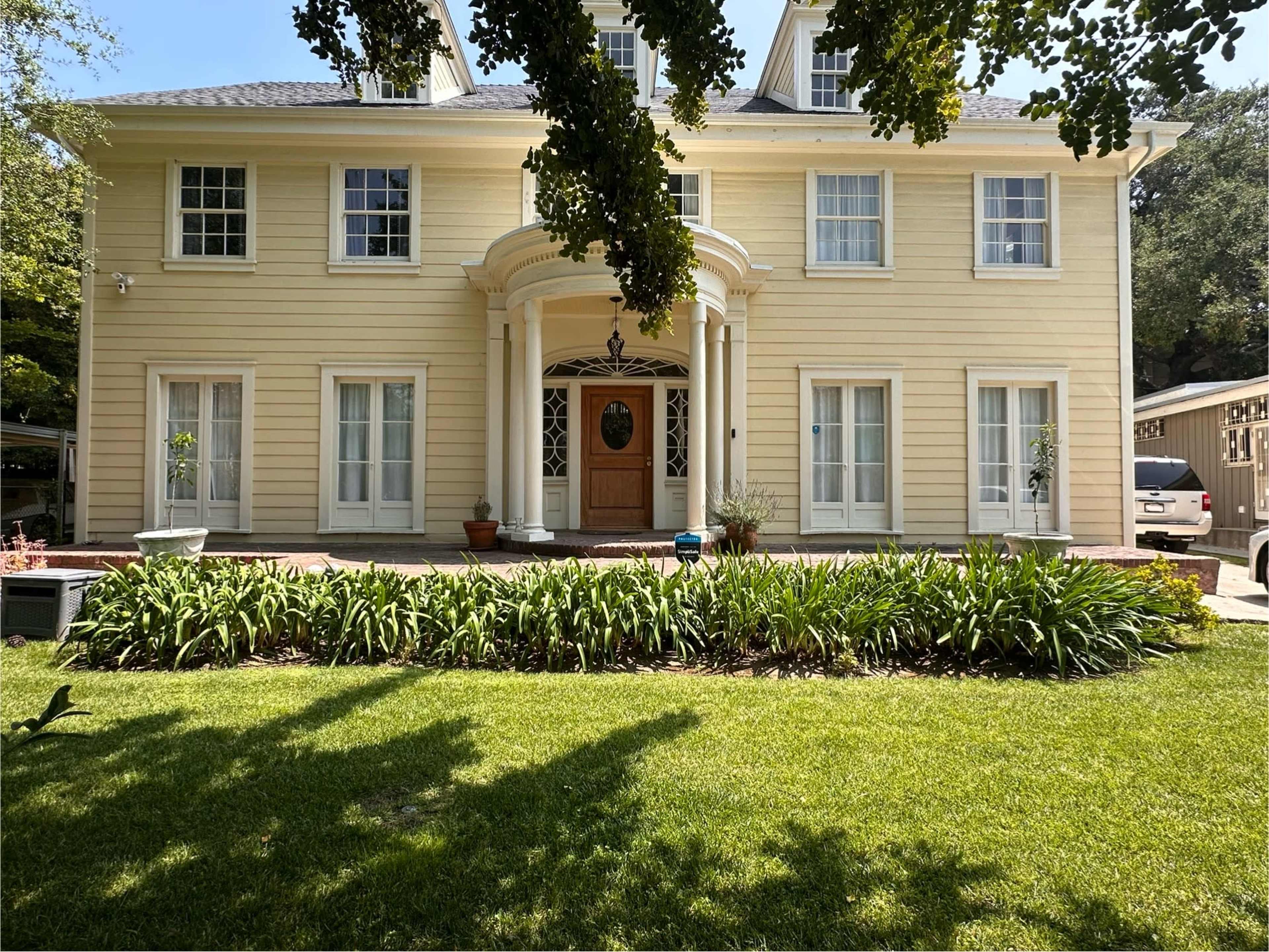 A two-story yellow house with white trim, featuring a central entrance and a landscaped front yard with green shrubs.