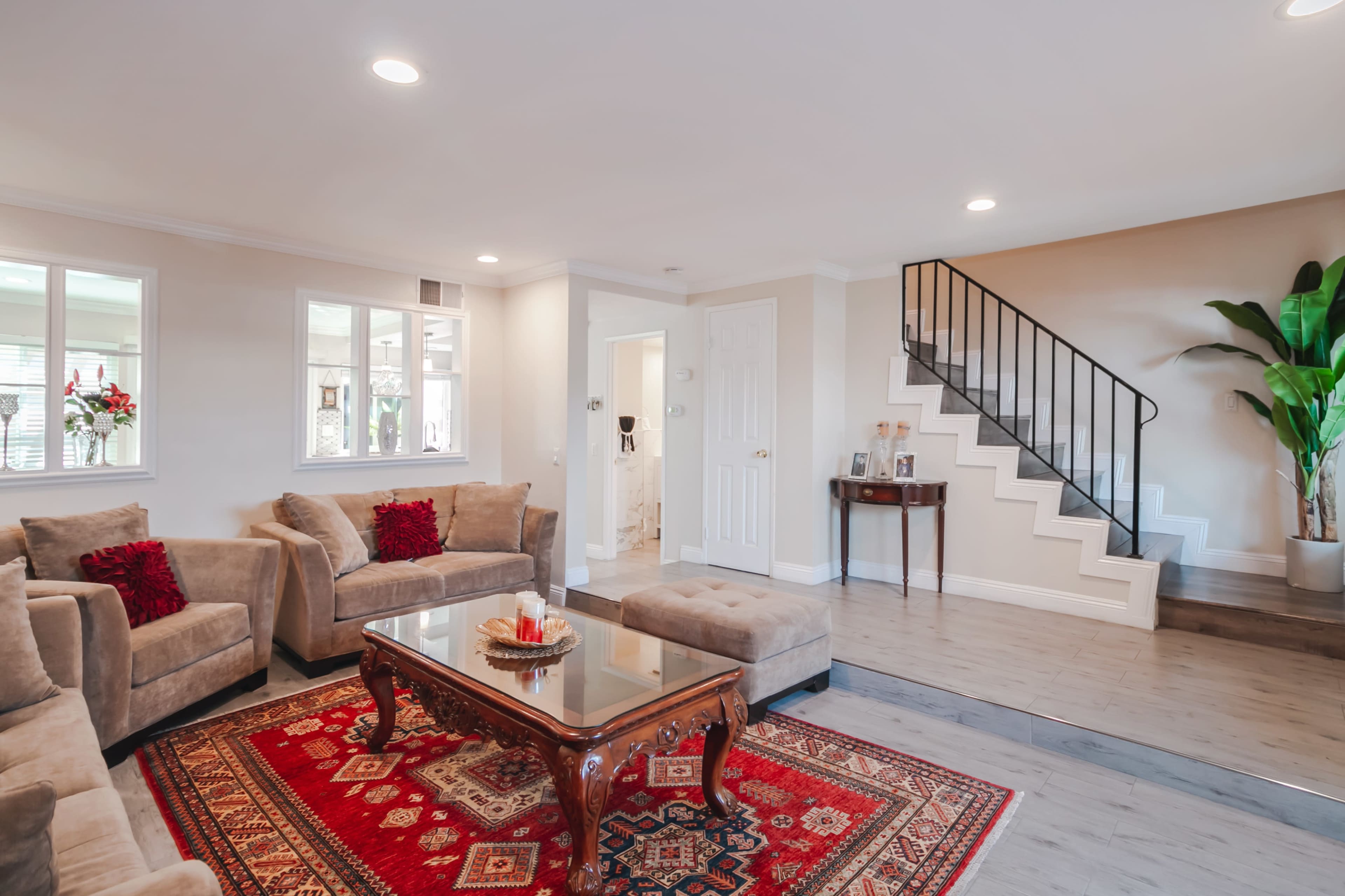 A living room with beige sofas, a glass coffee table on a decorative rug, and access to a staircase in the background.