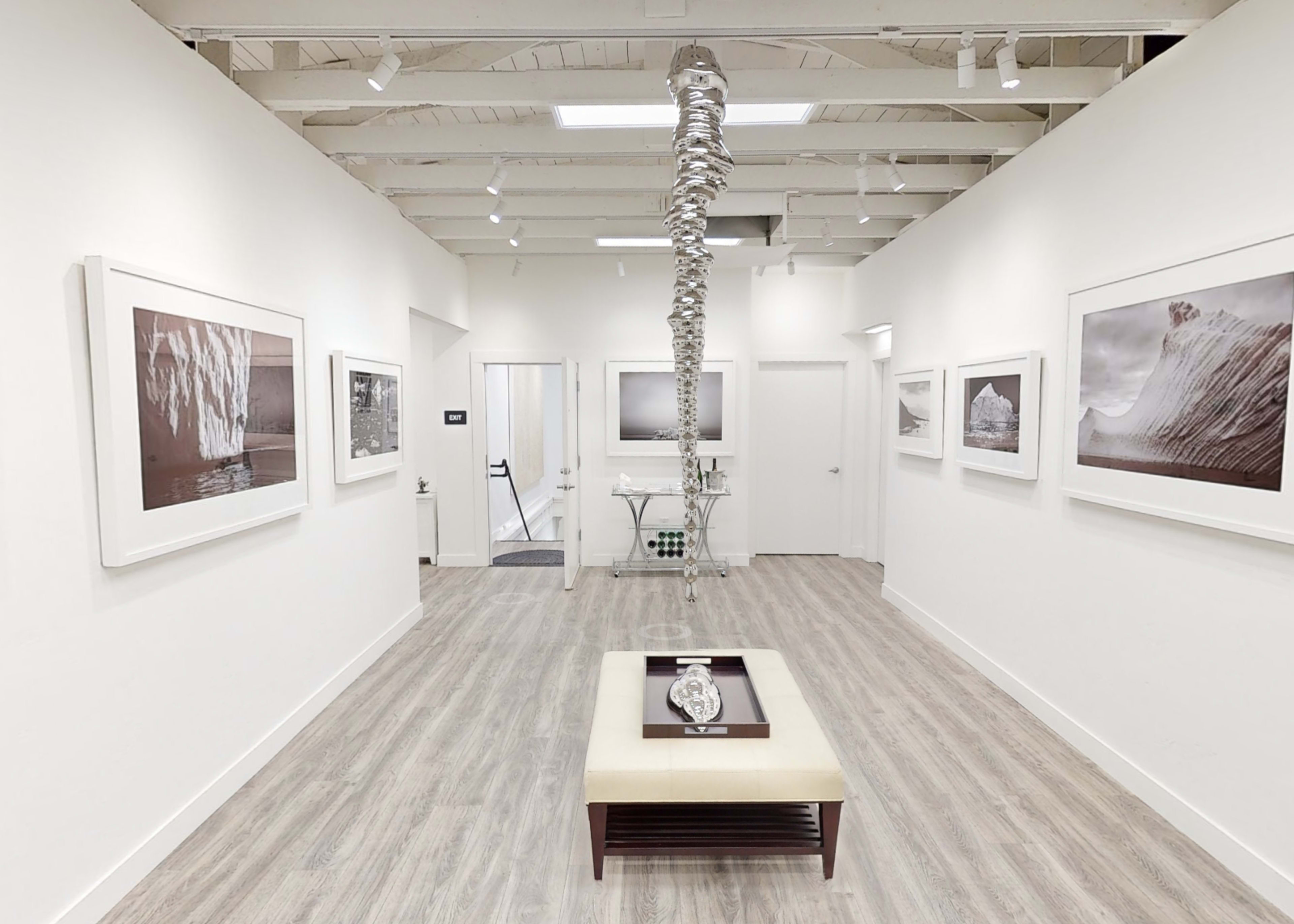 A modern art gallery with framed photographs on the walls, a central coffee table, and a sculptural installation hanging from the ceiling.