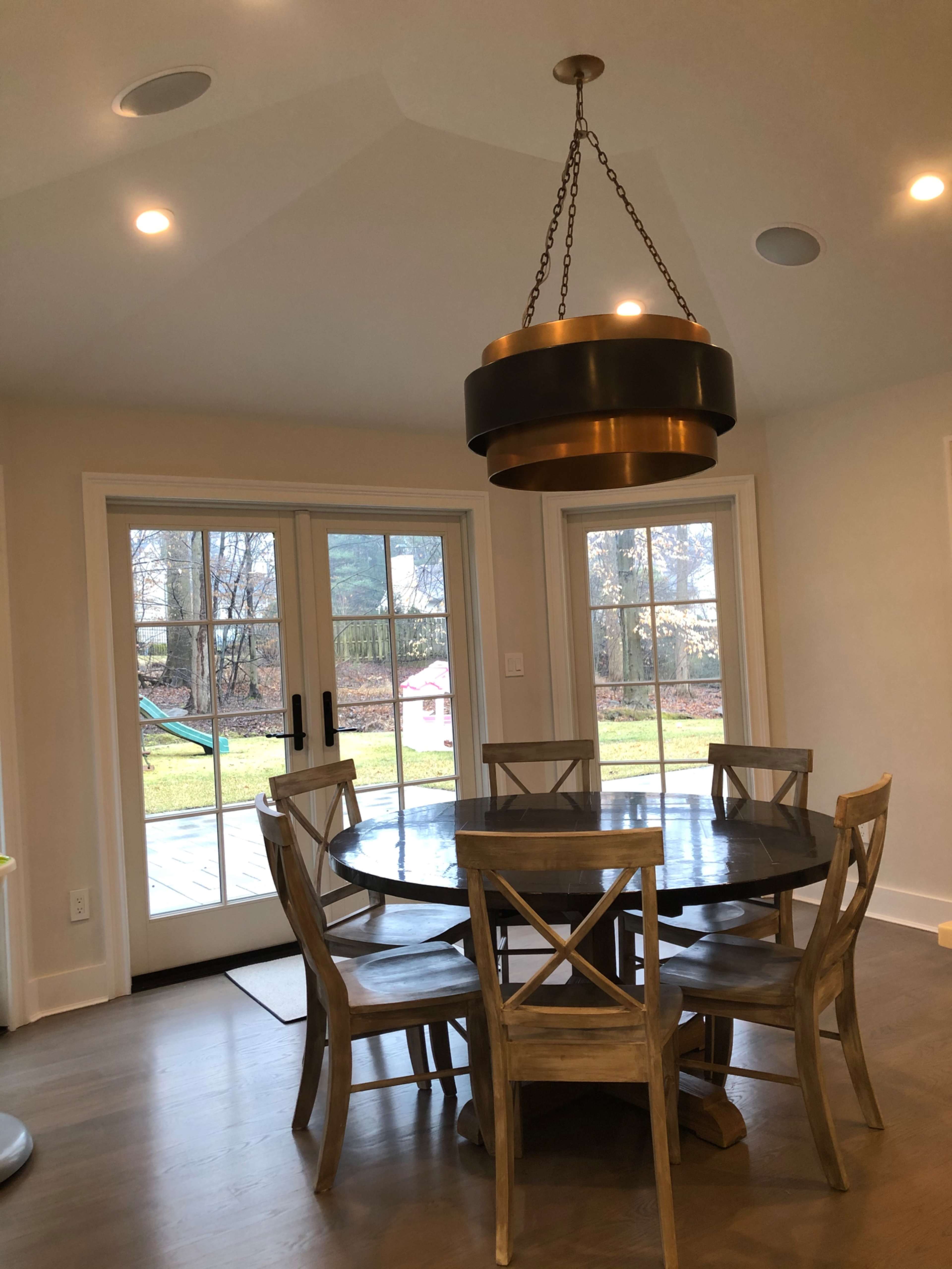 A round wooden dining table surrounded by six chairs is illuminated by a large pendant light, with sliding glass doors leading to a backyard in the background.