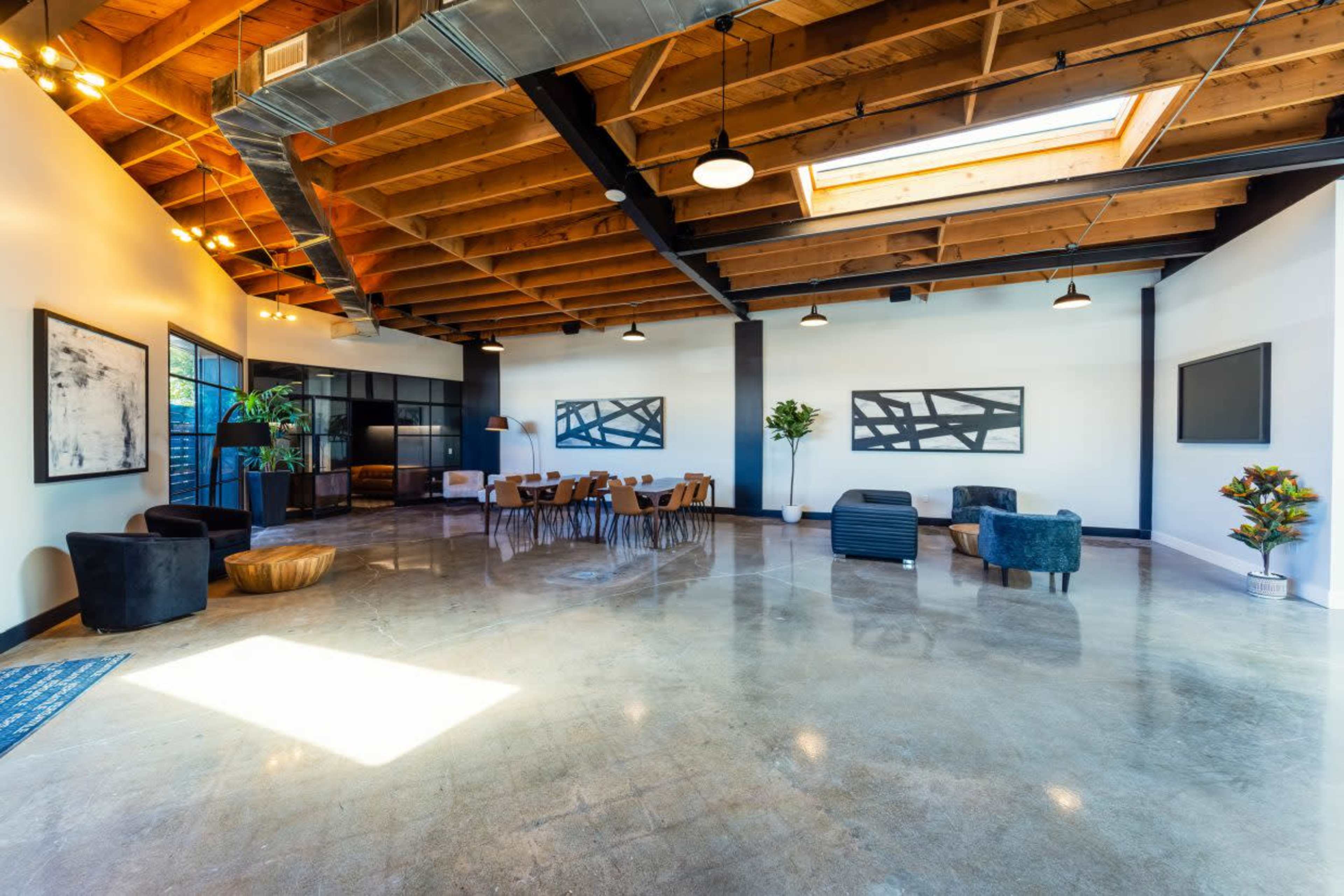 The image depicts a spacious, modern interior with high wooden ceilings, polished concrete flooring, several seating areas, and large windows allowing natural light to enter.