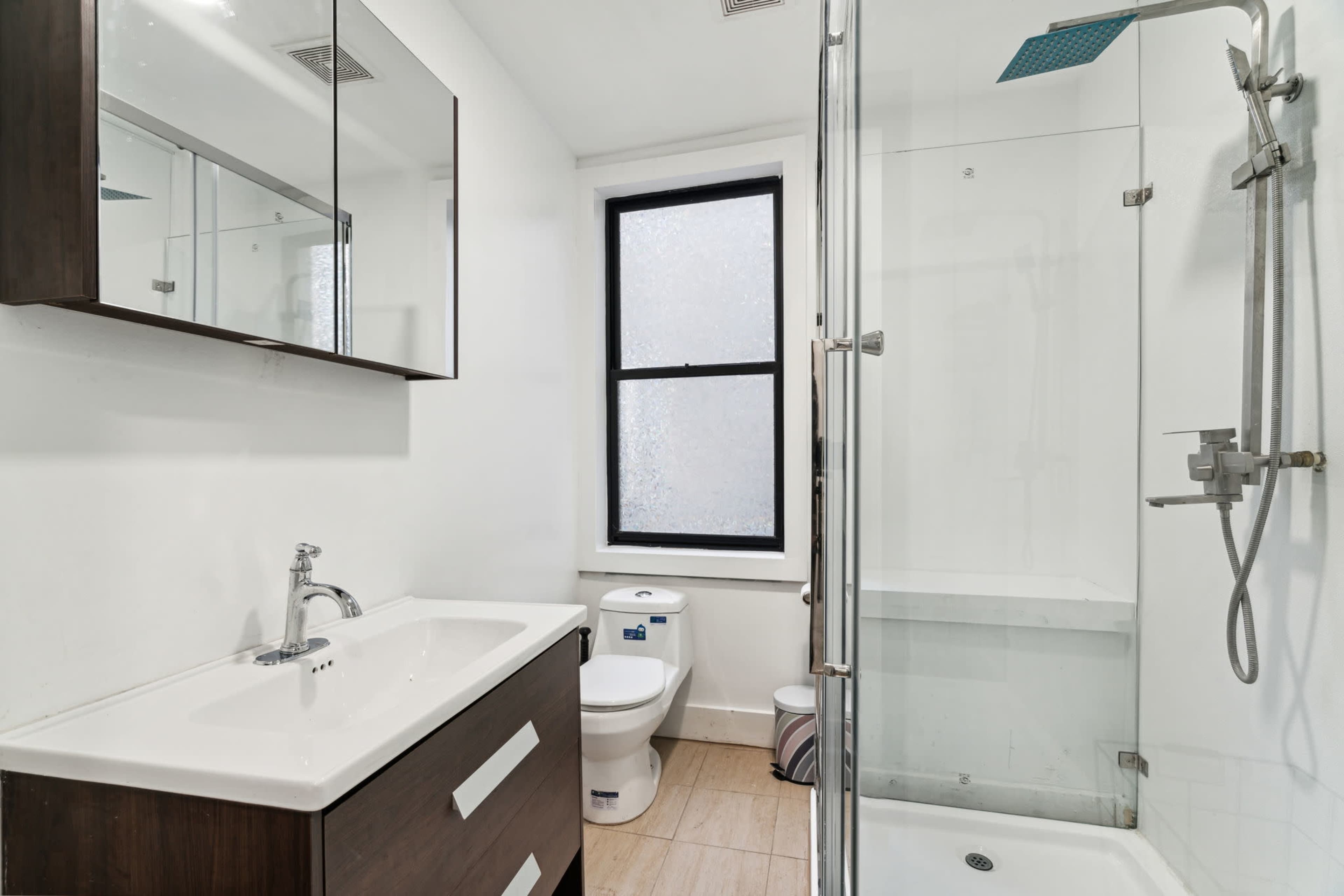 A modern bathroom featuring a shower with a glass enclosure, a wall-mounted mirror, a sink with cabinetry underneath, and a toilet beside a frosted window.
