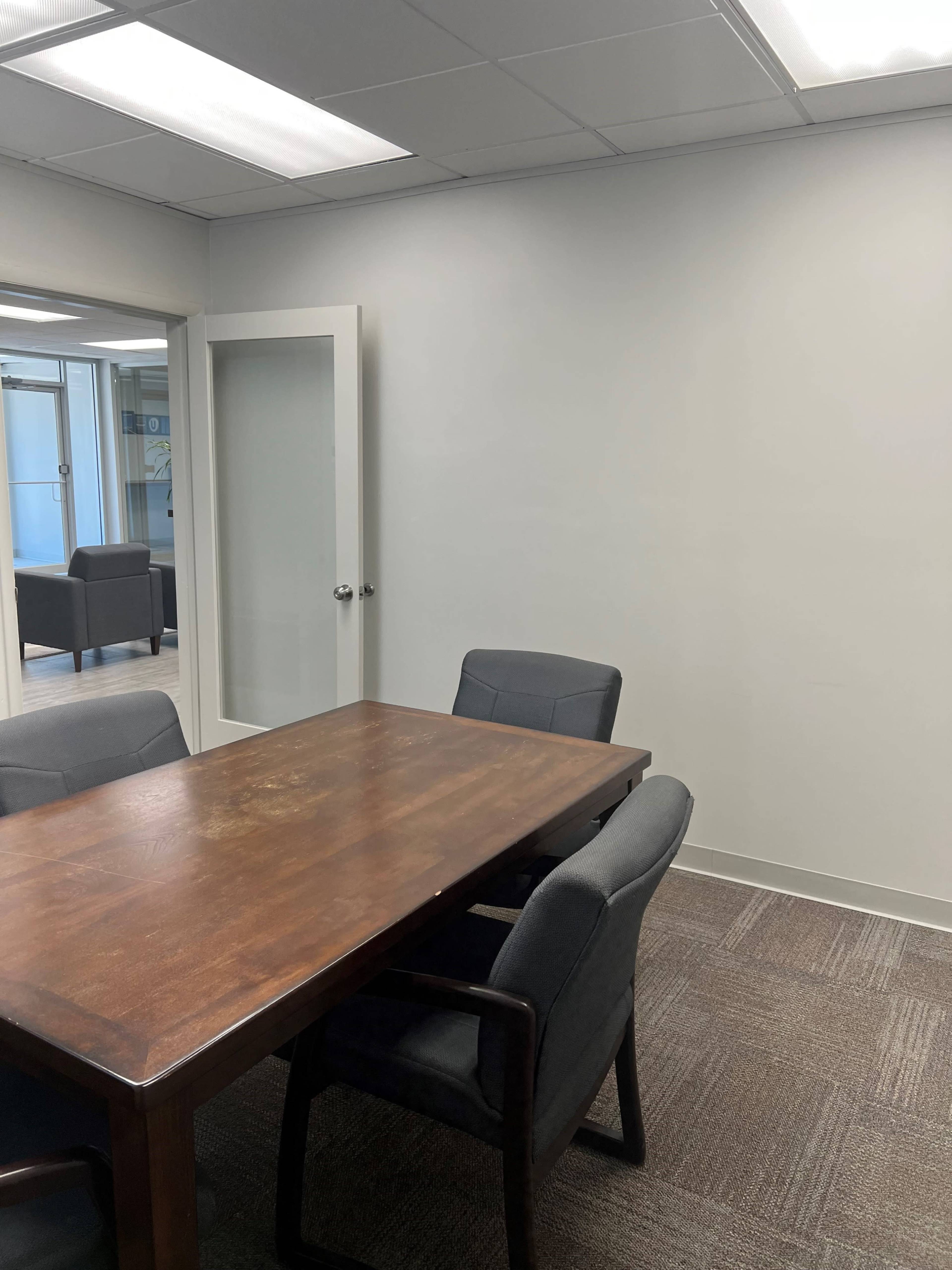 The image shows a conference room with a wooden table and four gray chairs, along with a partially open glass door leading to another space.